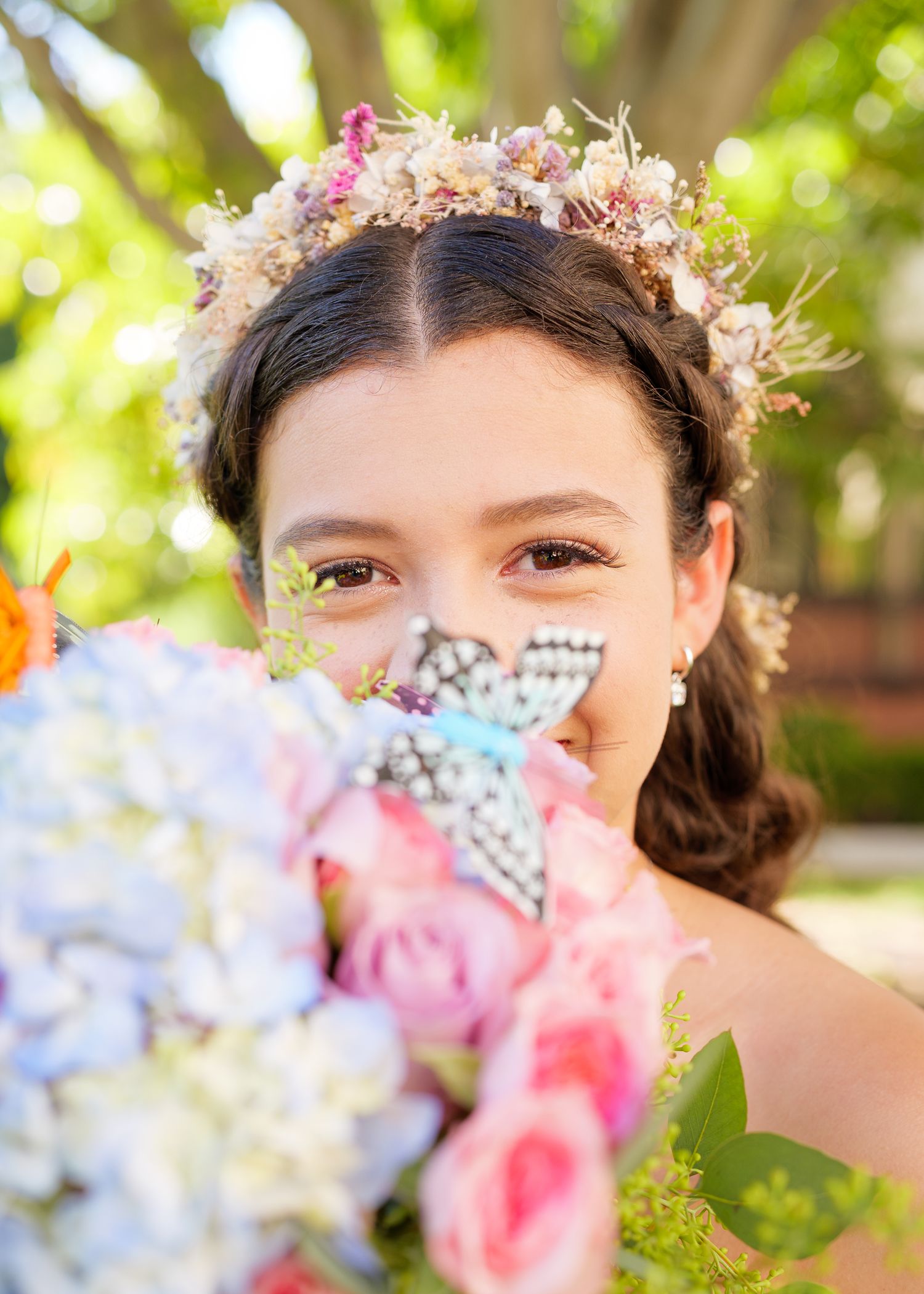 A delicate floral crown adorns the hair while holding a bouquet of pink roses and blue hydrangeas in soft natural light.
