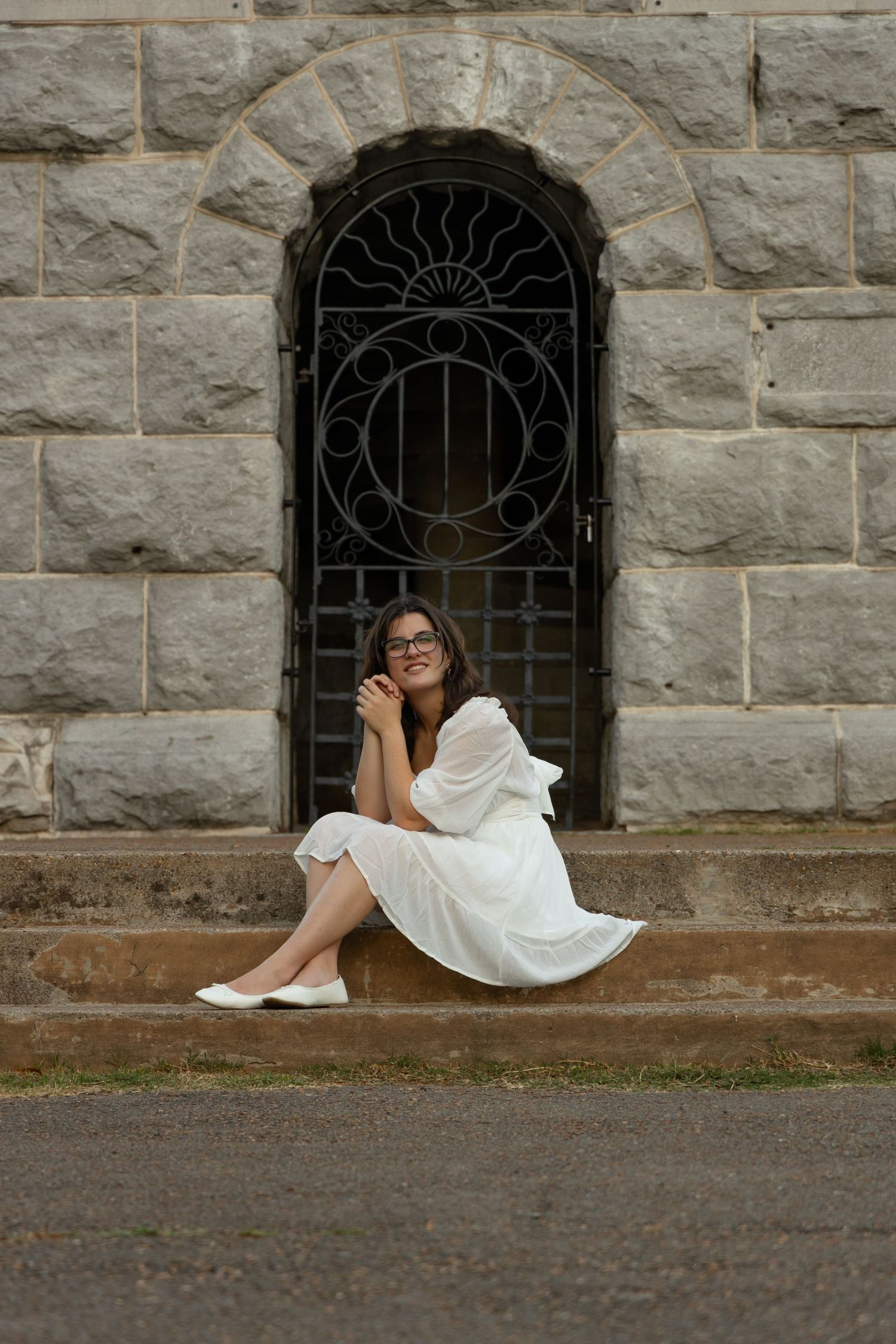 A woman in a white dress sits thoughtfully by an ornate iron gate entrance.