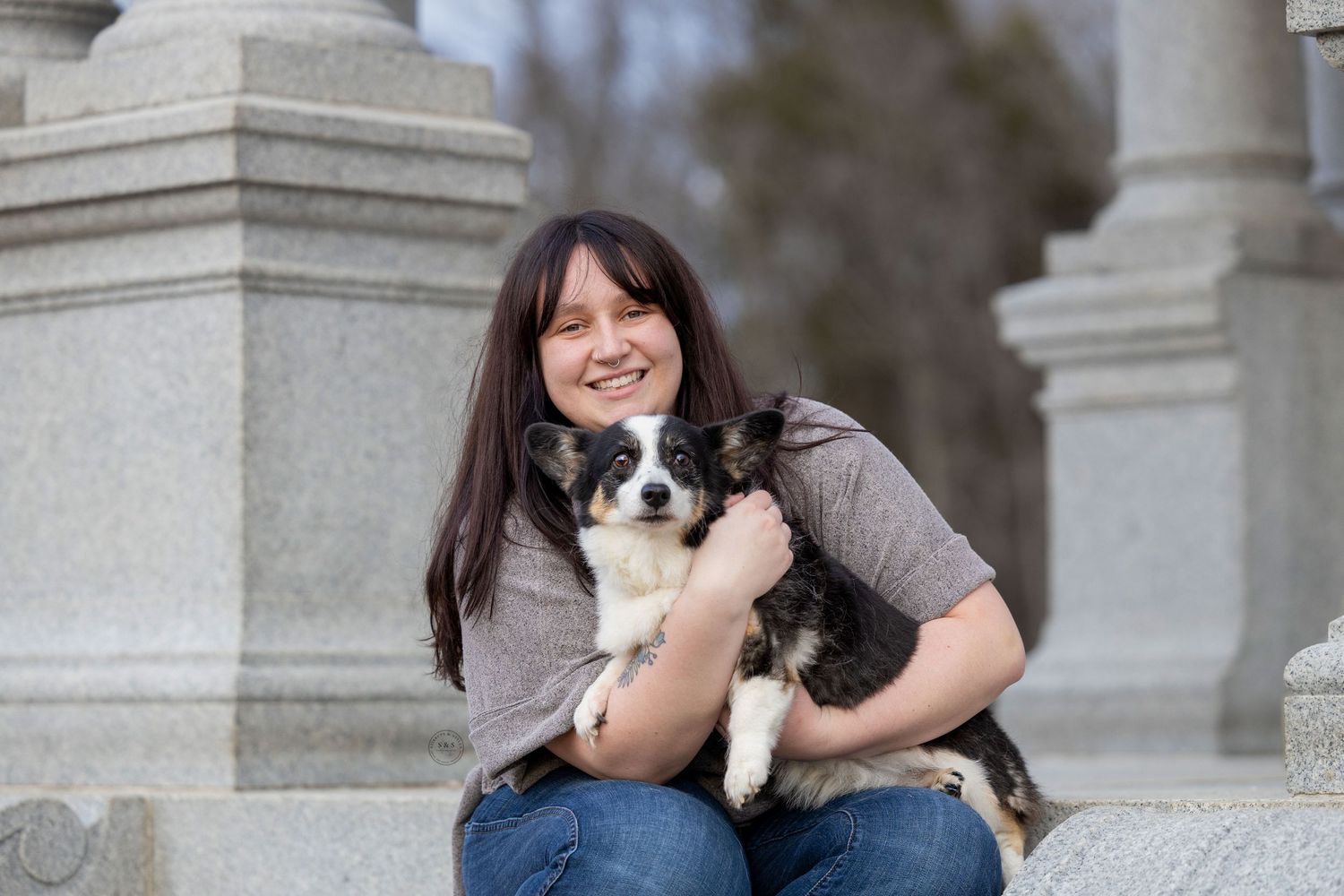 A happy person in a grey sweater sits on stone steps while holding a small black and white dog.