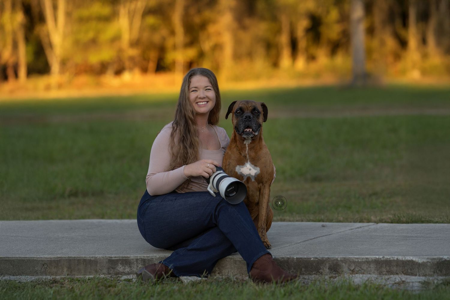 Woman smiling with boxer dog outdoors at golden hour, holding DSLR camera, seated on curb in park.