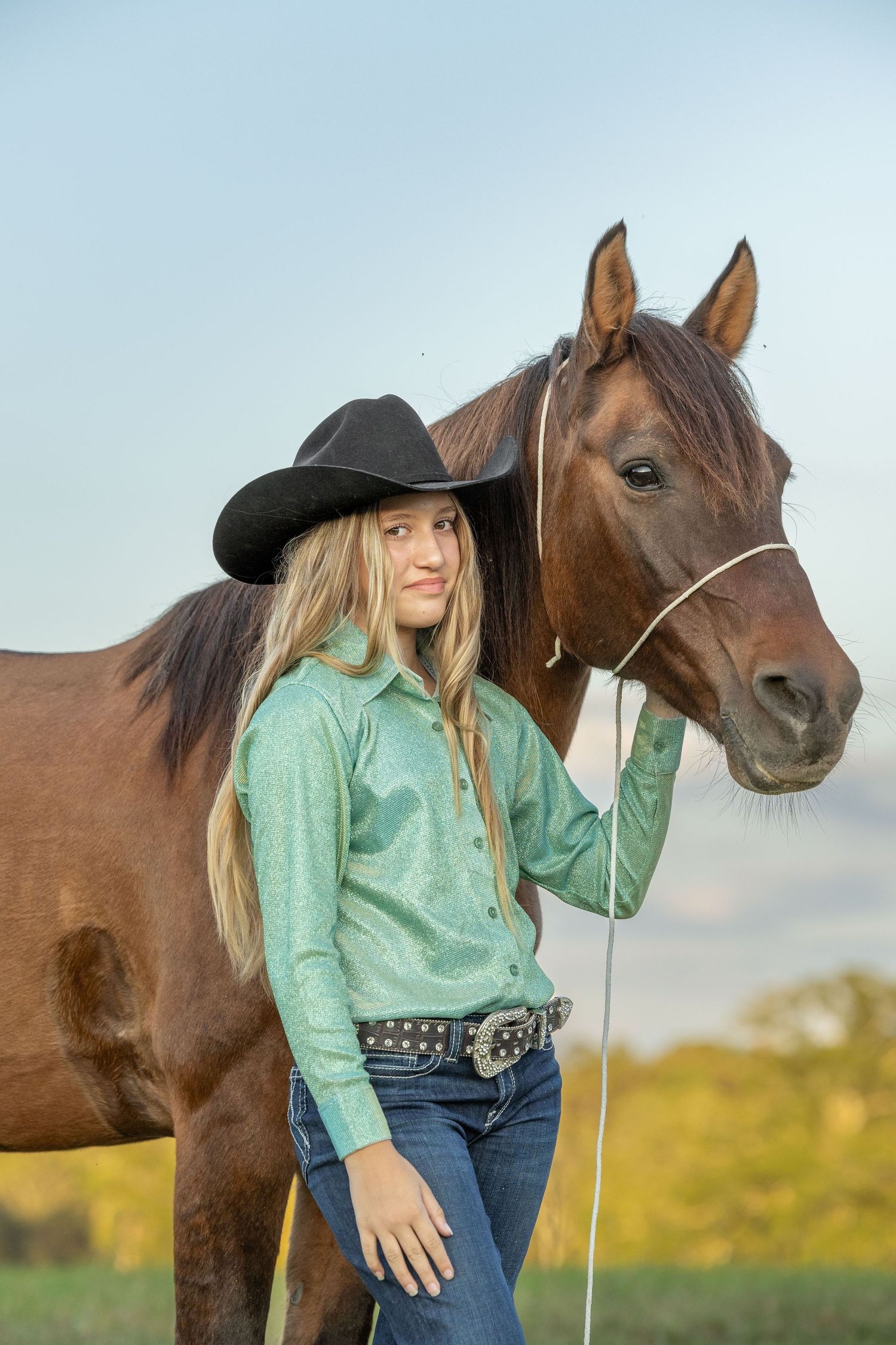 A woman in a mint green top poses with a brown horse in a golden field at sunset.