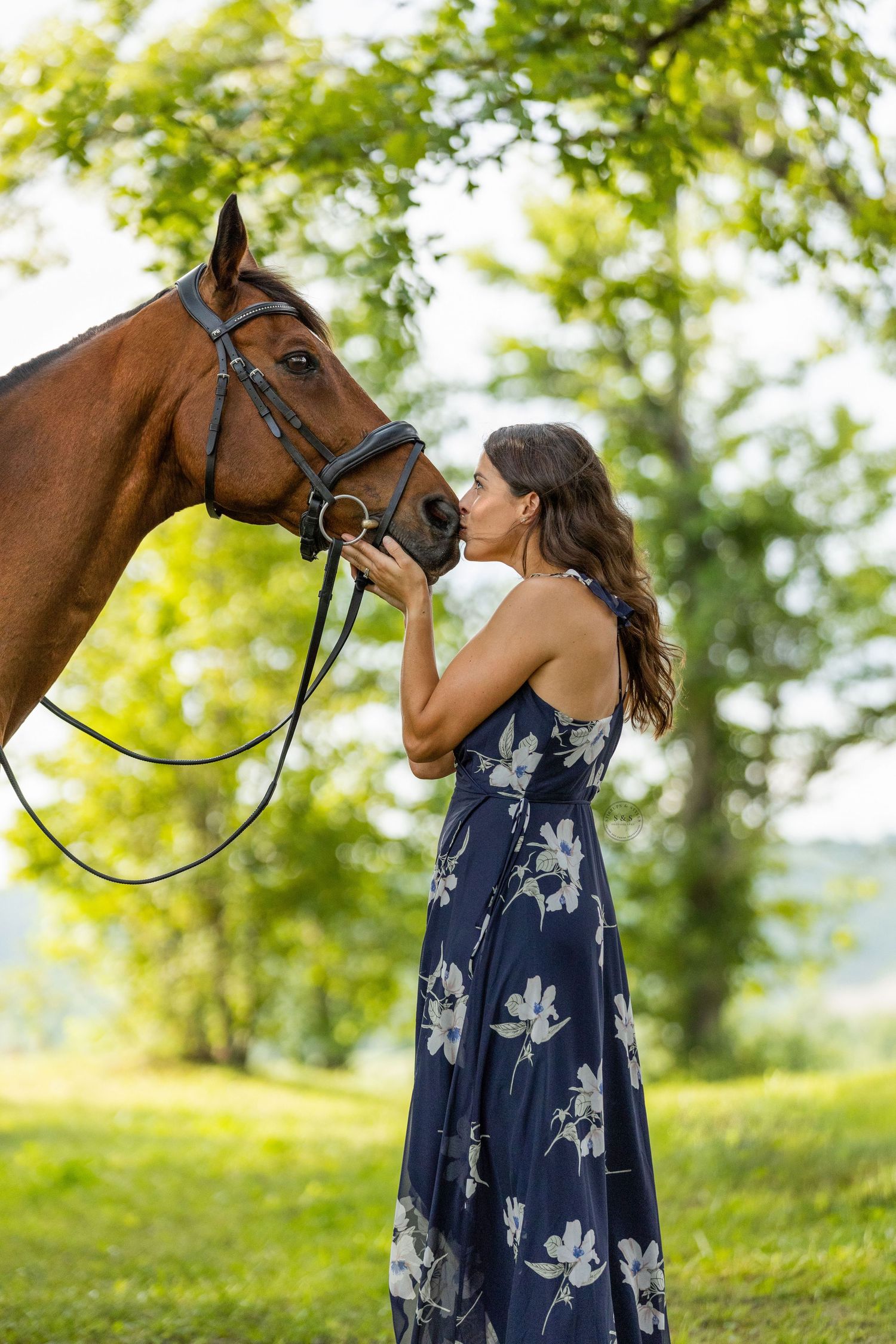 A tender moment captured between a bay horse and someone in a floral navy dress on a sunny summer day.