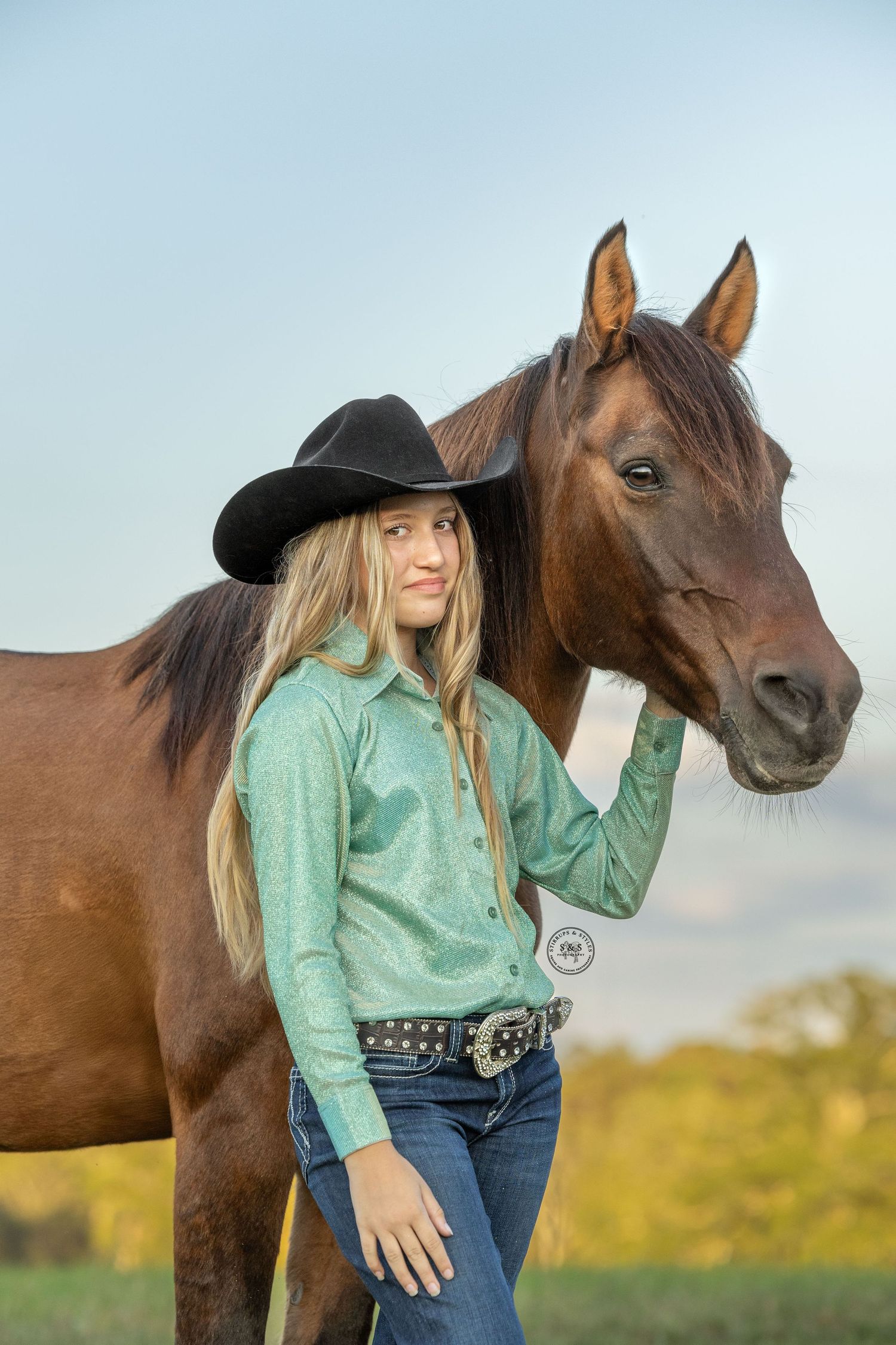 Woman in mint green top and cowboy hat poses with brown horse in golden sunlit field.