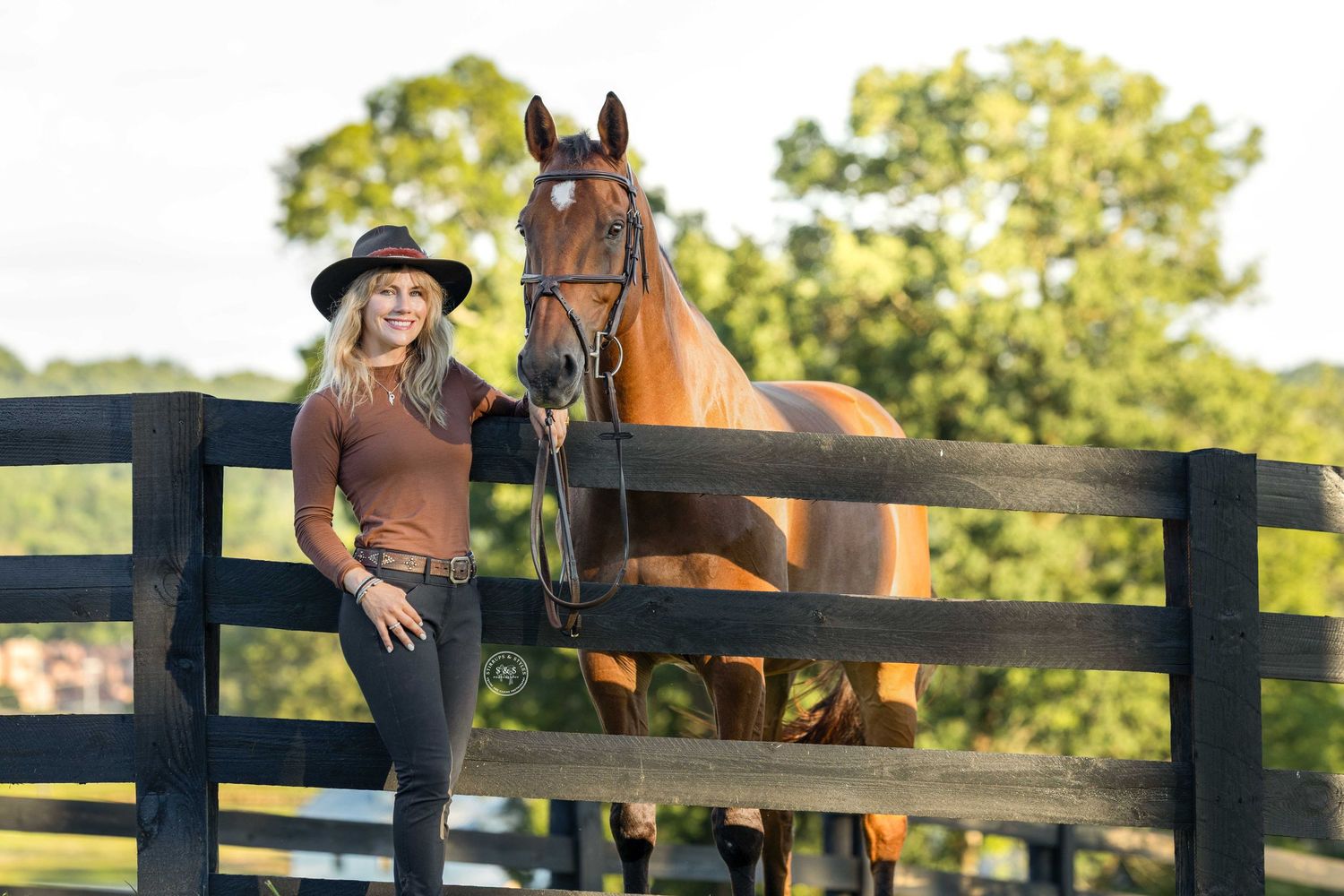 A pastoral scene shows horses resting by a wooden fence while someone sits on the fence railing at sunset.