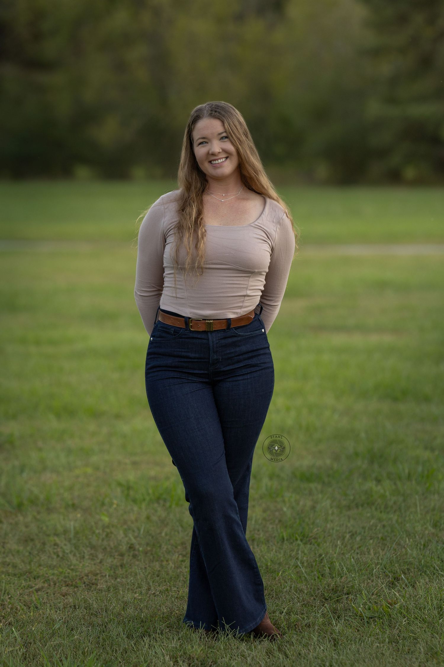 A person in a beige long-sleeve top and blue jeans poses casually on green grass in an outdoor setting.