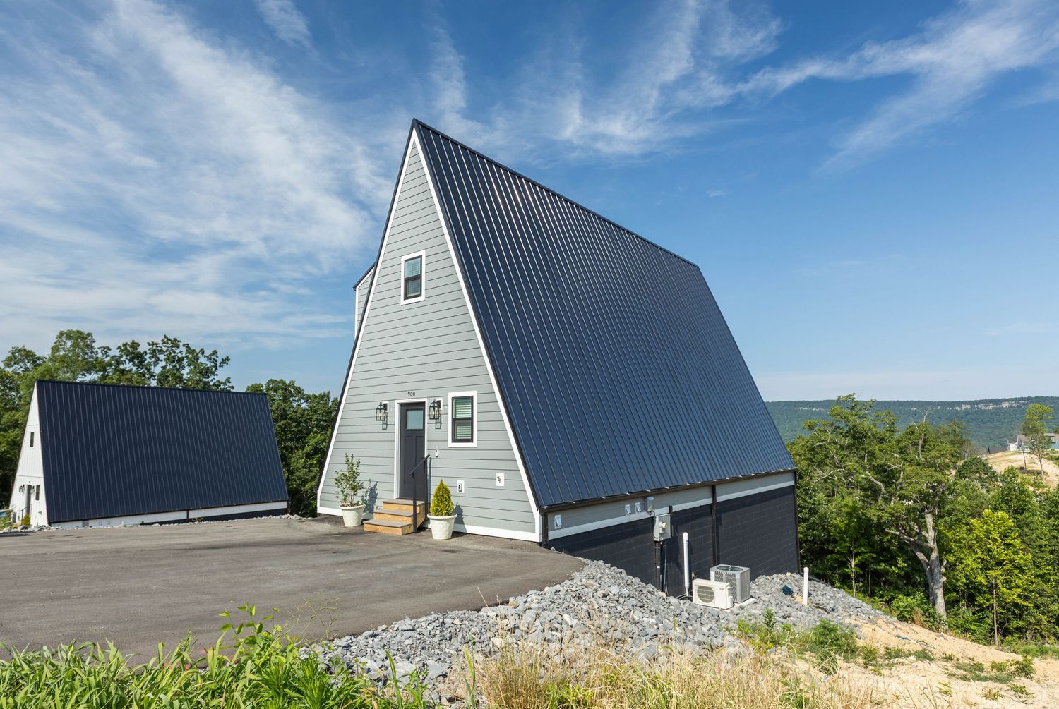 Modern A-frame vacation house with gray metal roof perched on hilltop overlooking green landscape on sunny day.