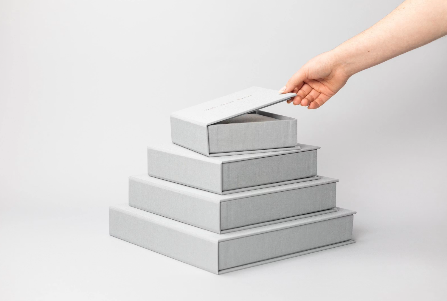 Hand lifting lid off stacked gray gift boxes arranged in pyramid shape on white background.