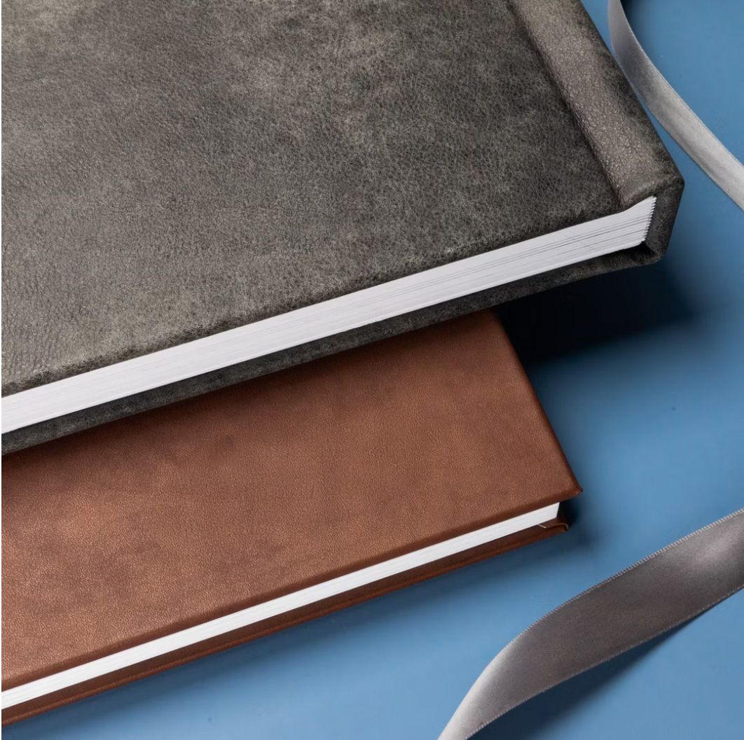 Two leather-bound hardcover notebooks in gray and brown displayed on a blue surface with a silver ribbon.