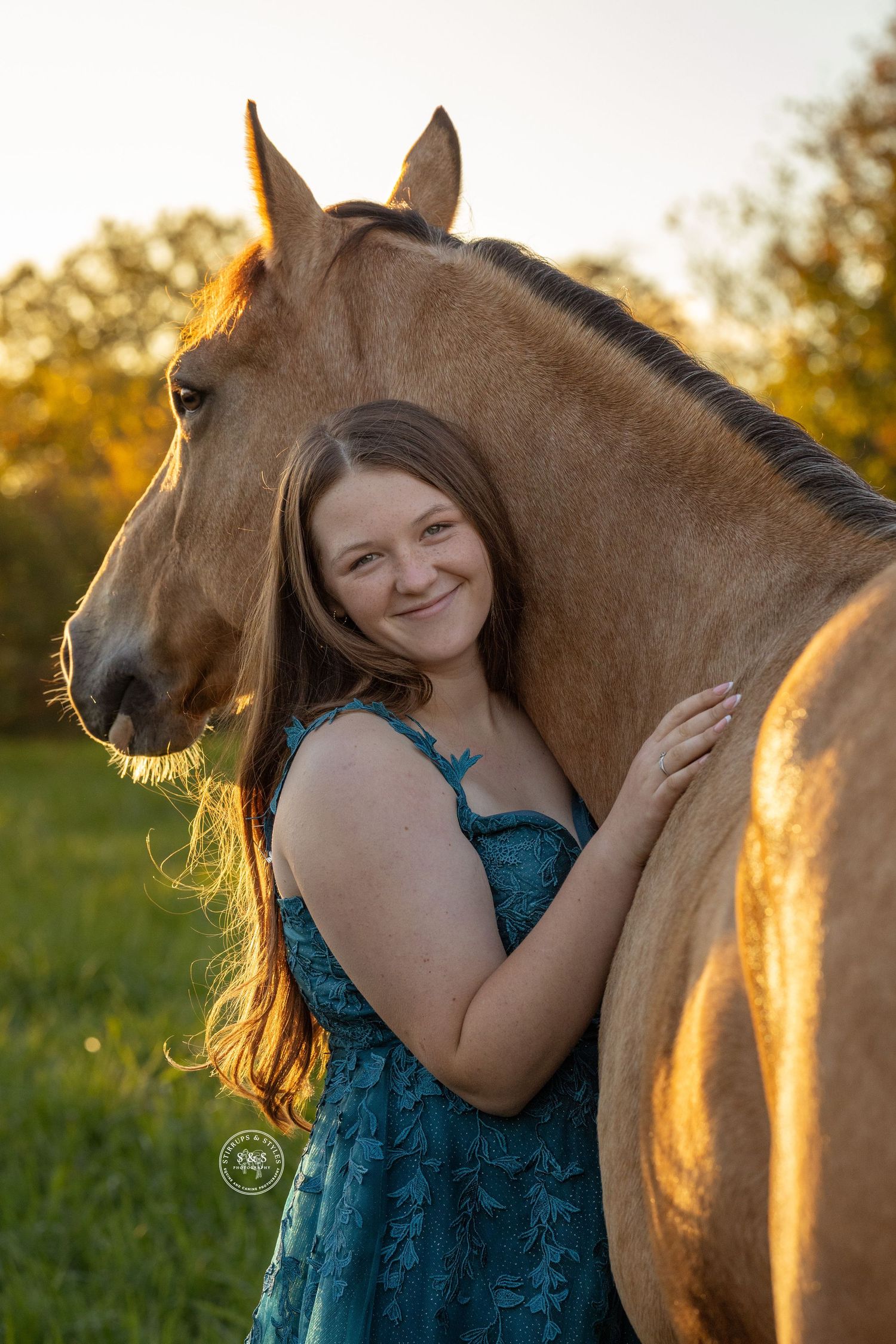 A person in a teal dress stands next to a brown horse in a sunlit field during golden hour.