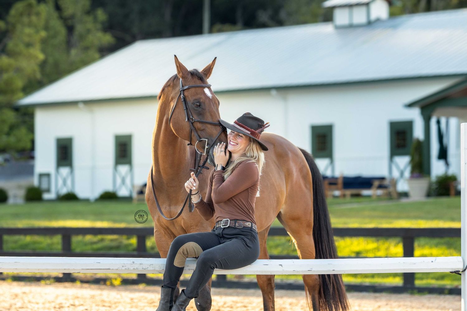 A rider sits on a chestnut horse in front of a white barn during a summer day.