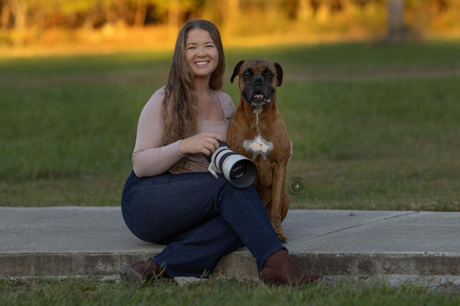 A photographer sits with their Boxer dog on concrete steps during golden hour while holding a camera.