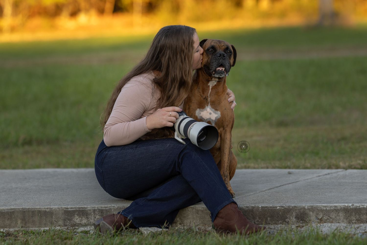 Photographer with camera crouches next to a brown dog in a grassy field at sunset.