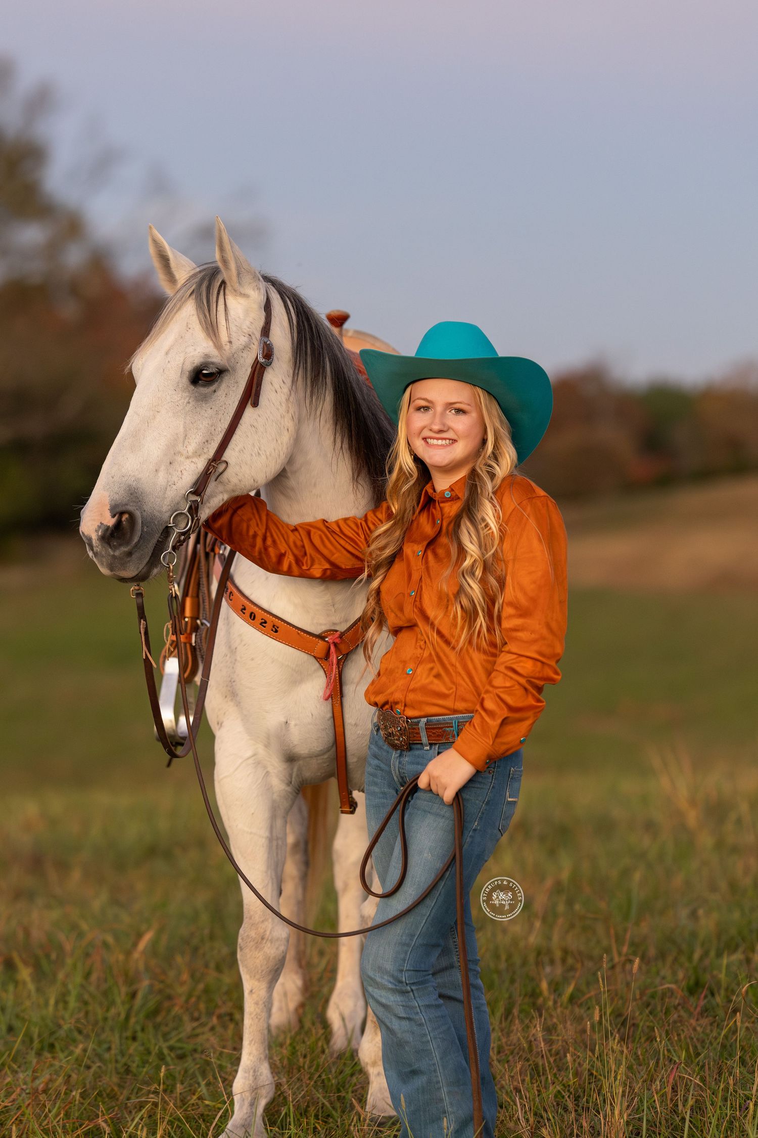 A beautiful white horse stands in a grassy field at sunset with someone wearing an orange jacket and teal hat.