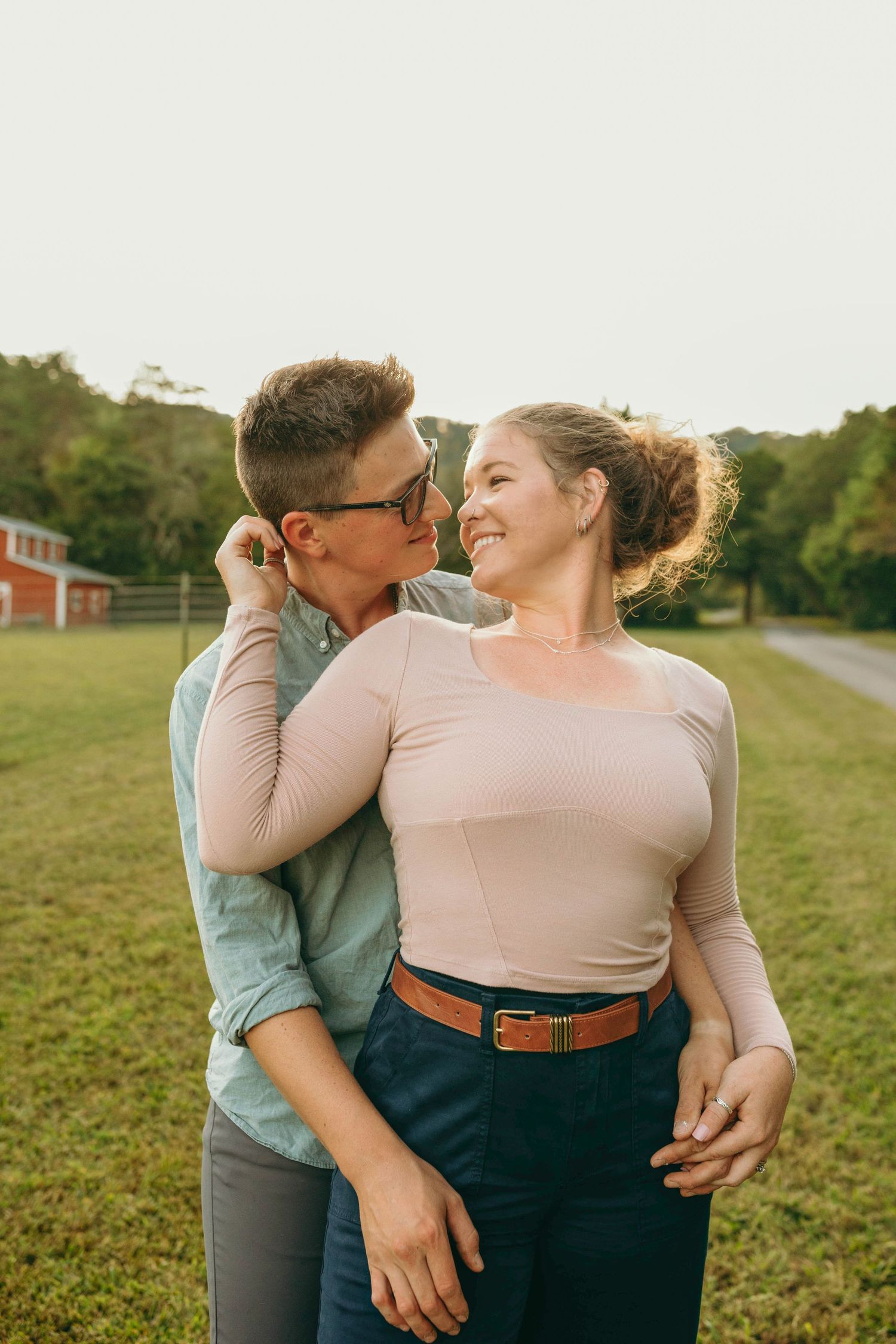 A couple embraces in a romantic moment against a pastoral farm field backdrop at golden hour.