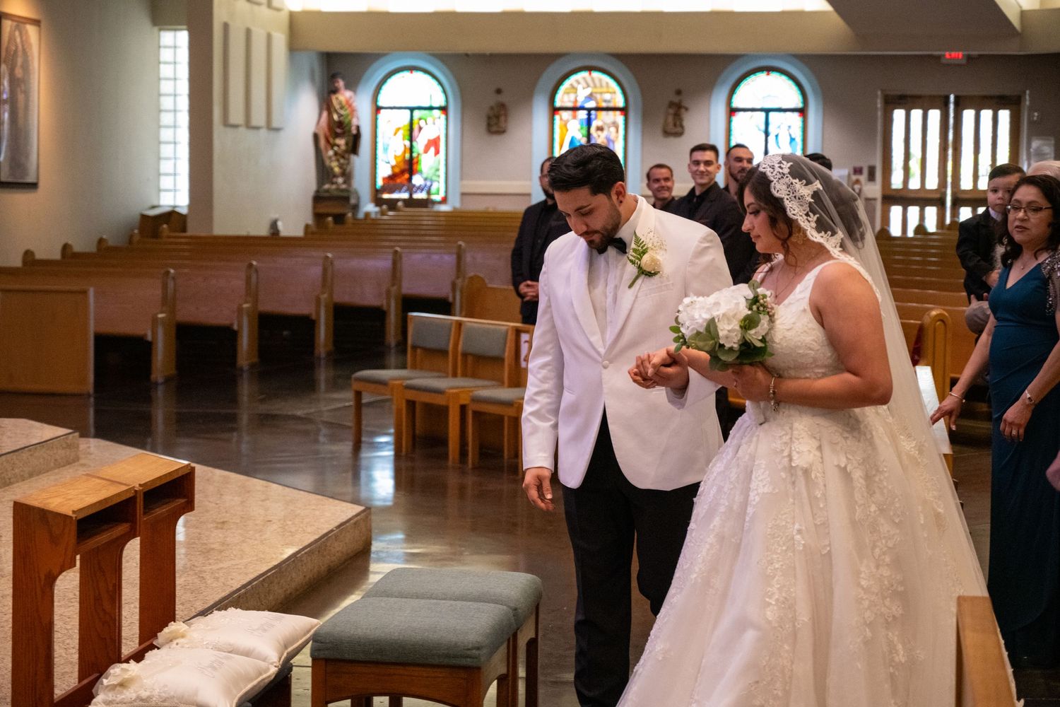A couple stands together in a church during their wedding ceremony wearing a white dress and pink tuxedo jacket.