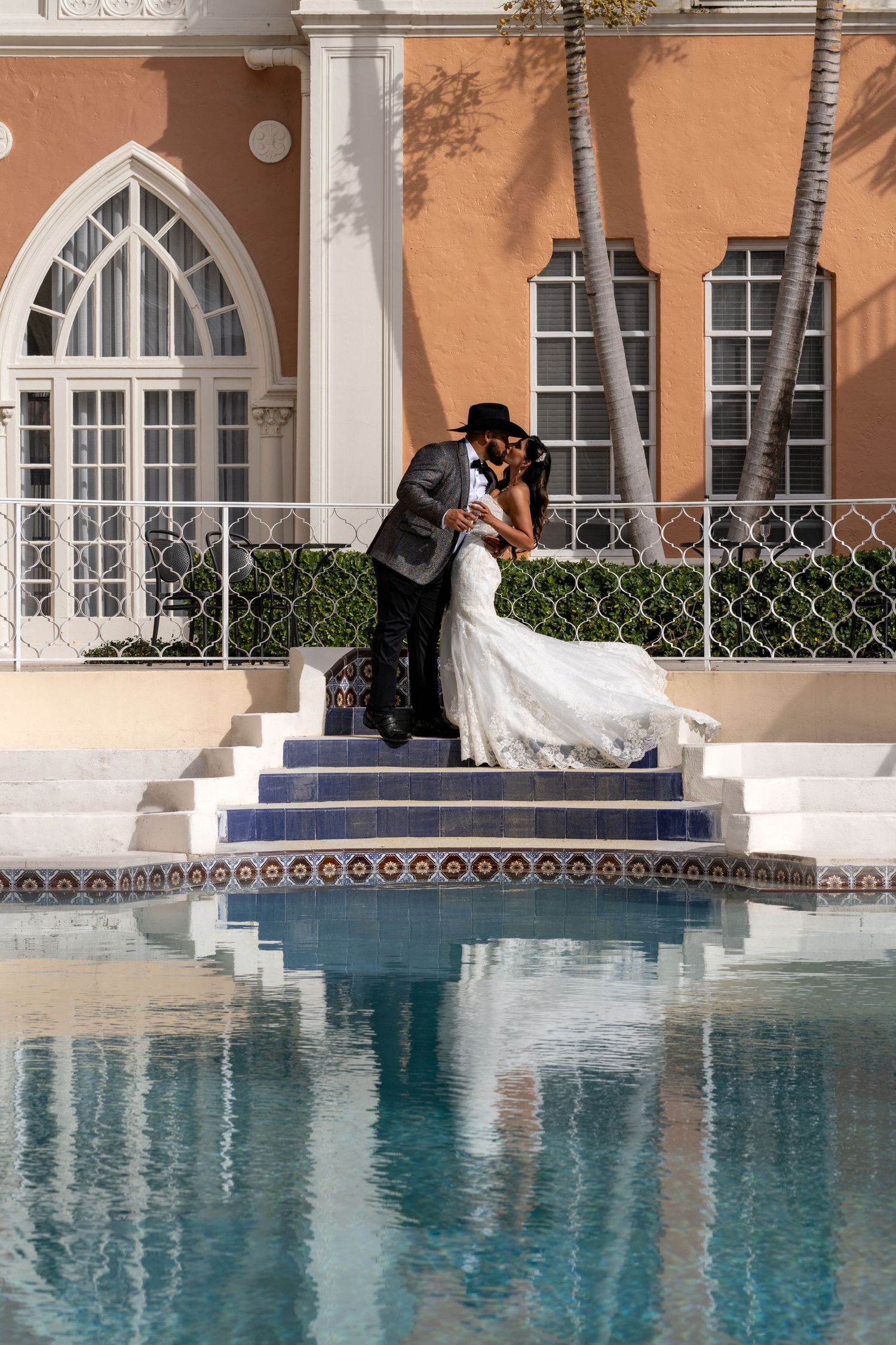 A couple embraces on elegant tiled steps beside a reflective pool at a Mediterranean-style venue with arched doorways and palm trees.