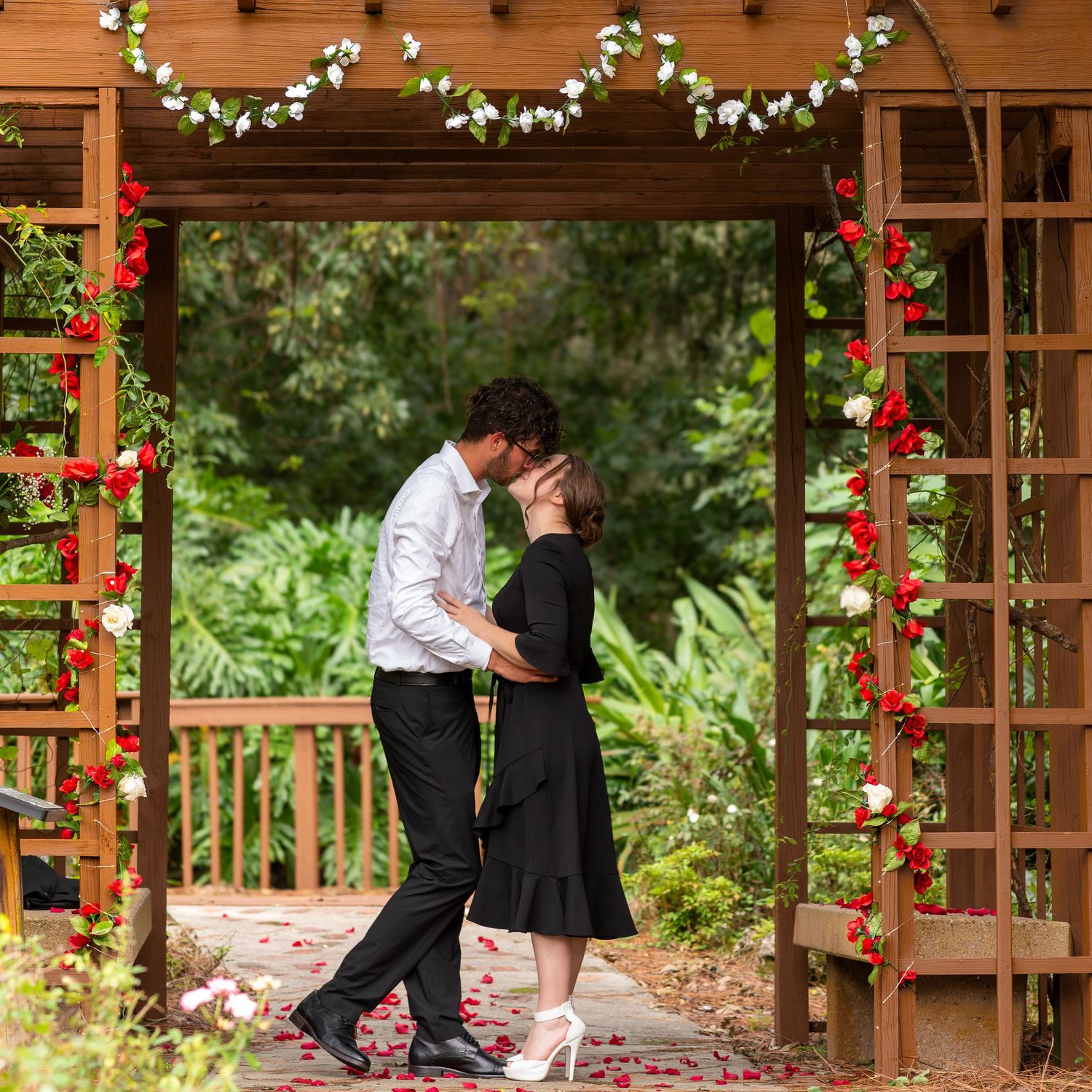 Romantic couple sharing an intimate moment under a wooden pergola decorated with floral garlands and red roses.