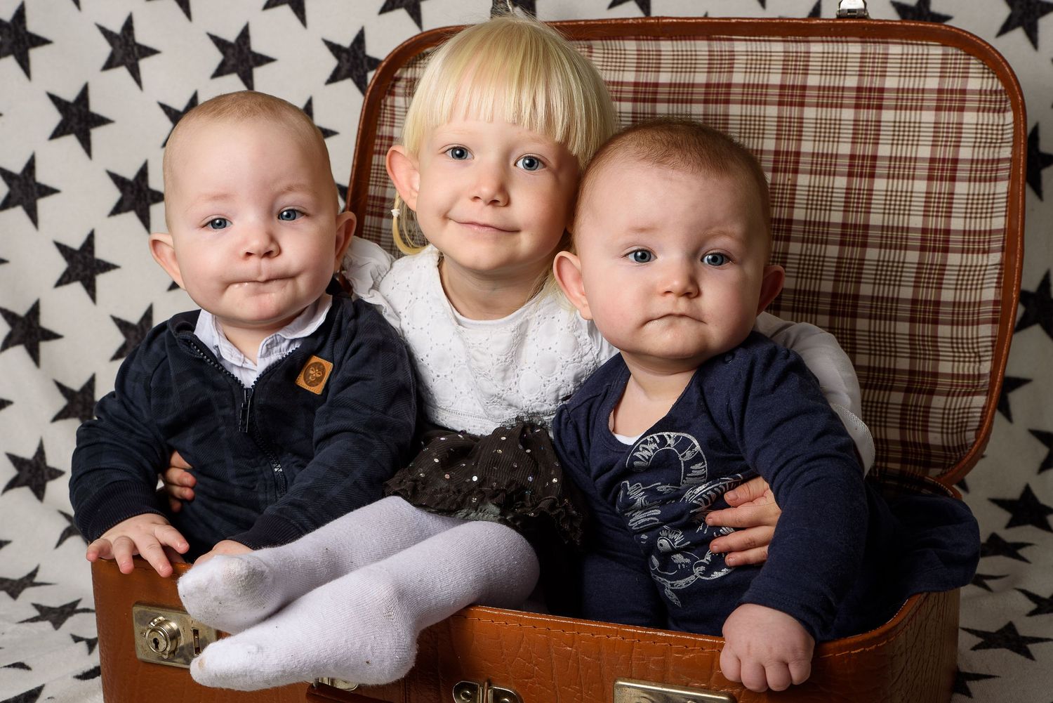 Three young children, two babies and a toddler, sitting together in a vintage brown suitcase against a star-patterned backdrop.