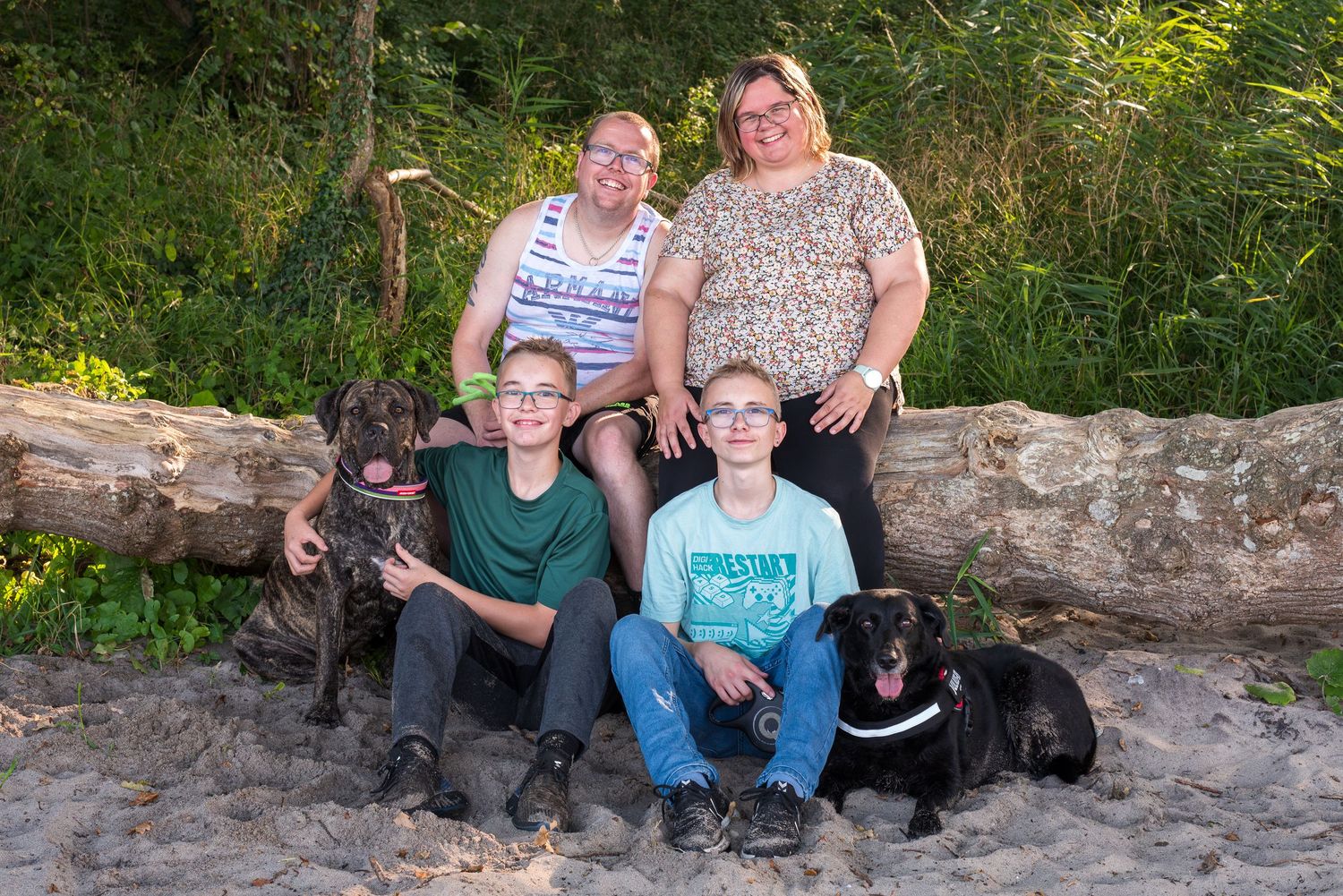 Family portrait with two adults, two boys, and two black dogs sitting on sandy ground near a fallen log outdoors.