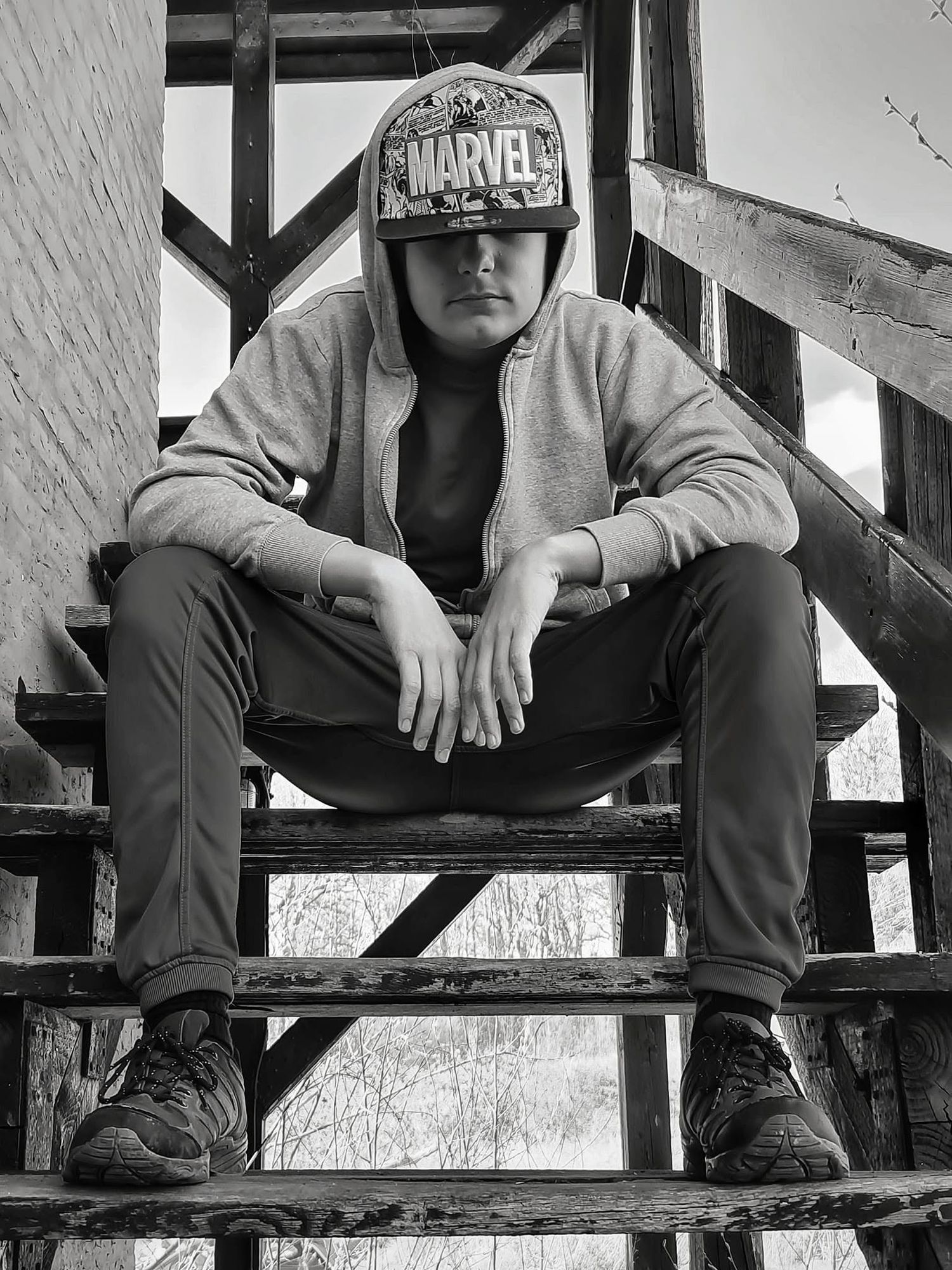Black and white photo of a teen in a Marvel cap and hoodie sitting on rustic wooden stairs outdoors.