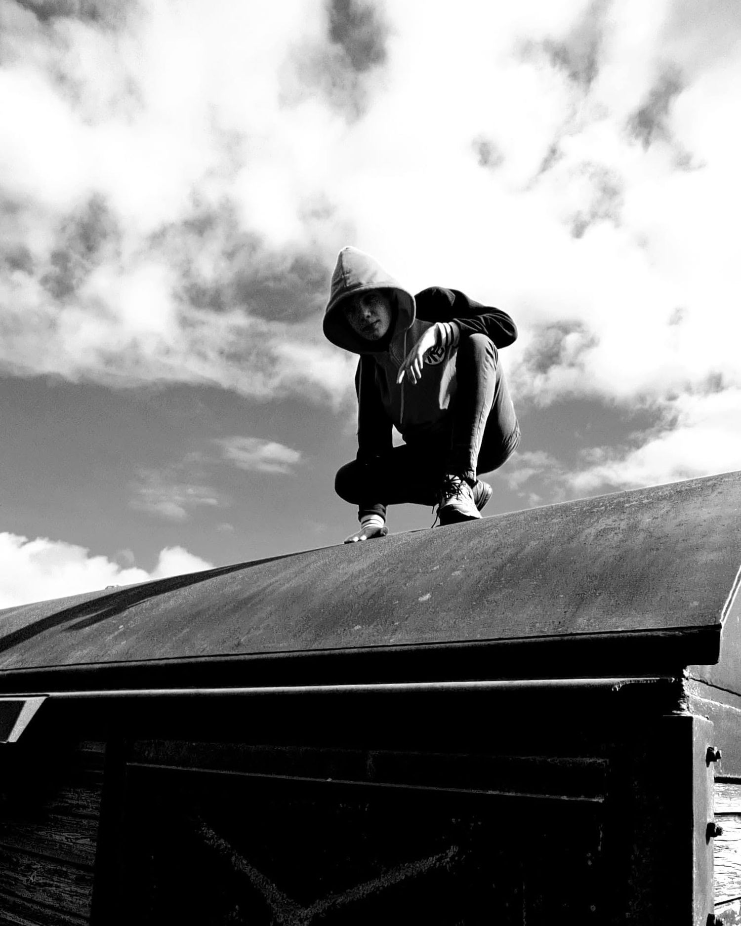 Person in hoodie crouching on rooftop against dramatic cloudy sky in black and white photo.