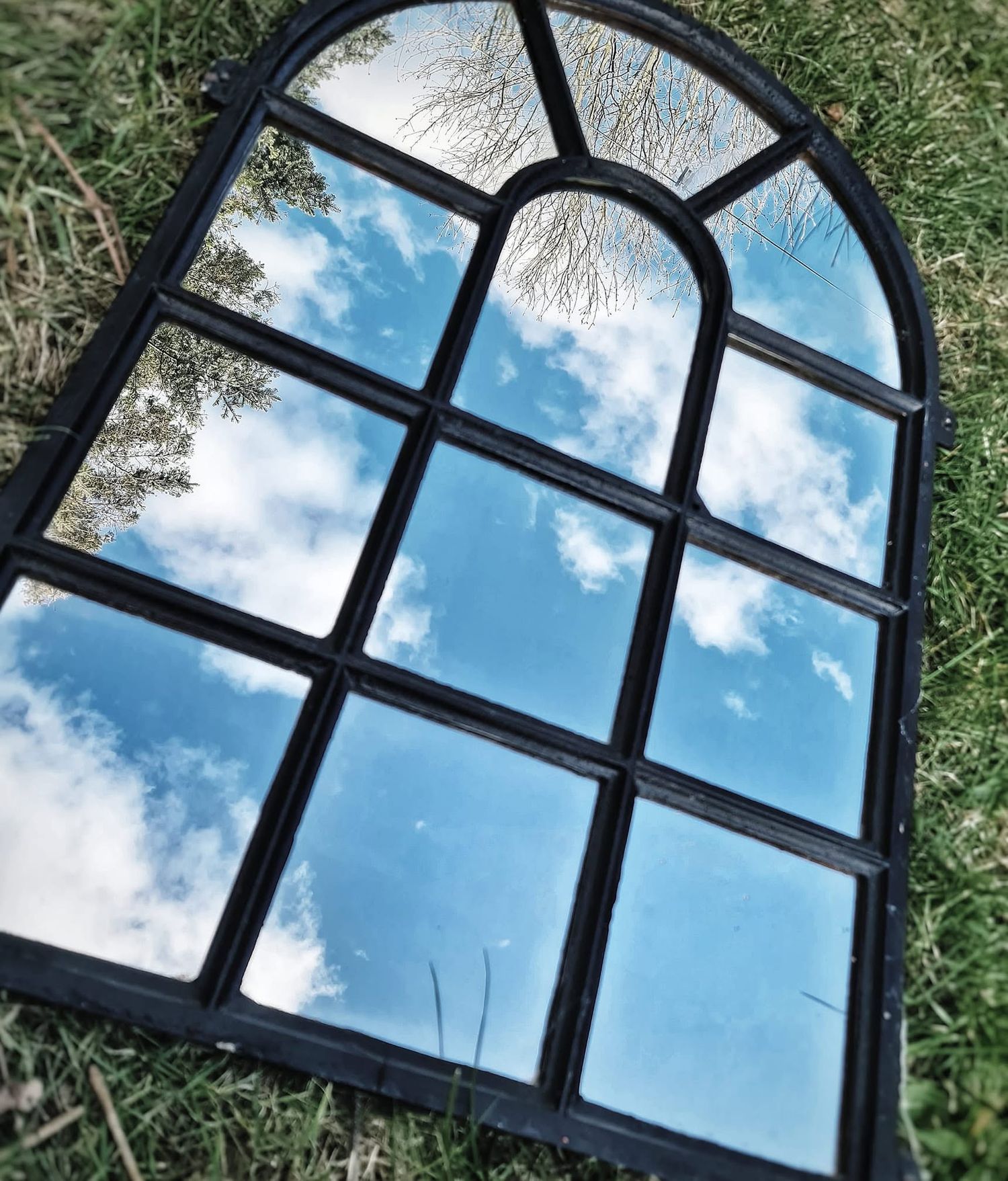 Arched window mirror lying on grass reflecting blue sky and clouds.
