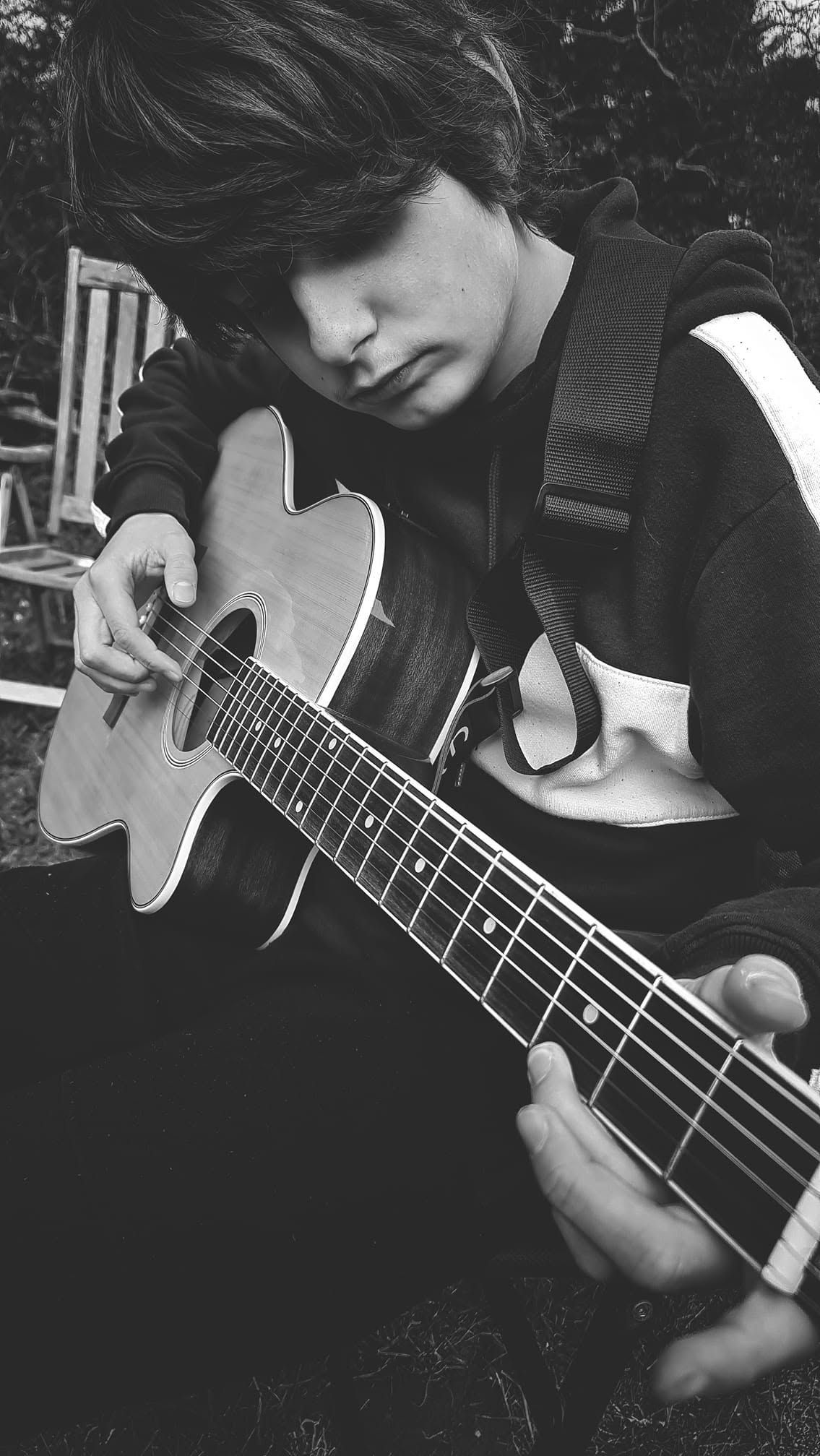 Black and white photo of a young man with dark hair playing an acoustic guitar outdoors.