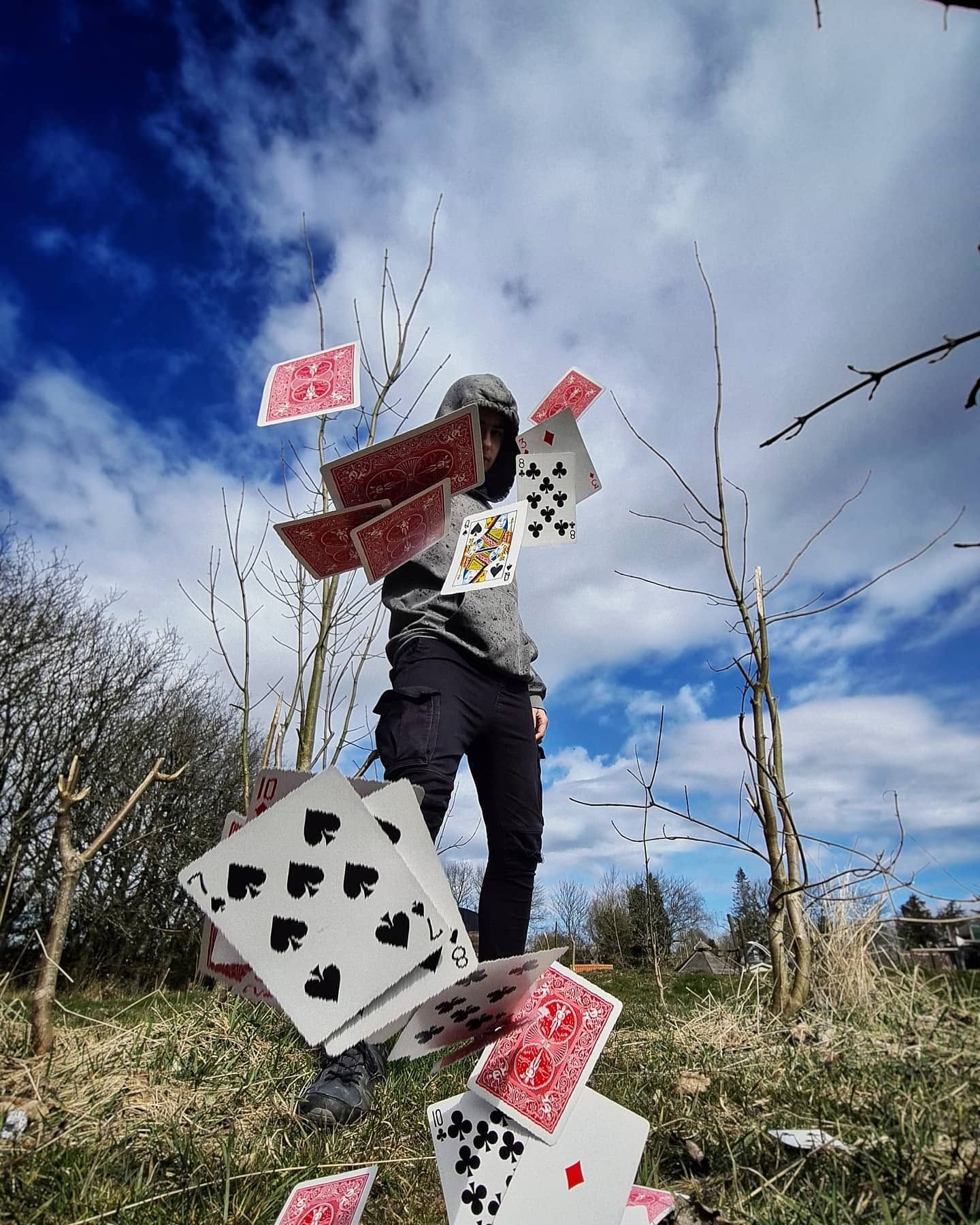 Person performing card magic trick outdoors with playing cards flying in the air against a cloudy blue sky.