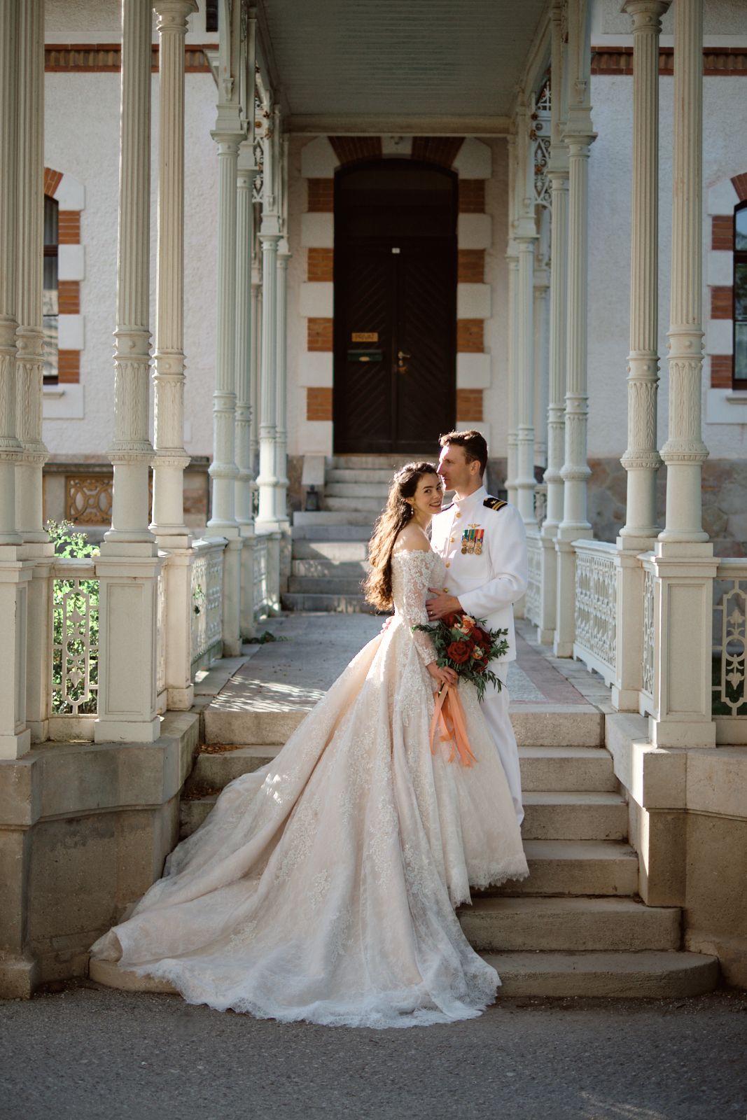 Bride in elegant wedding dress with train falling down steps, smiling at the camera. Groom wearing a navy uniform holding her. Wedding photo