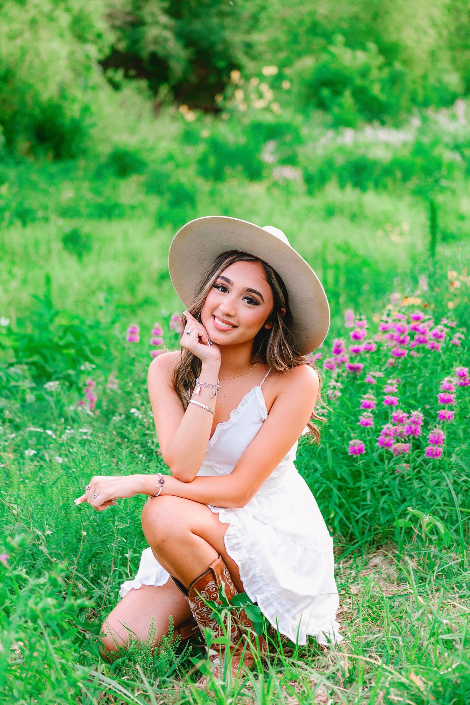 Senior girl in Houston, Texas kneeling in a field of flowers wearing a sun hat and white dress.