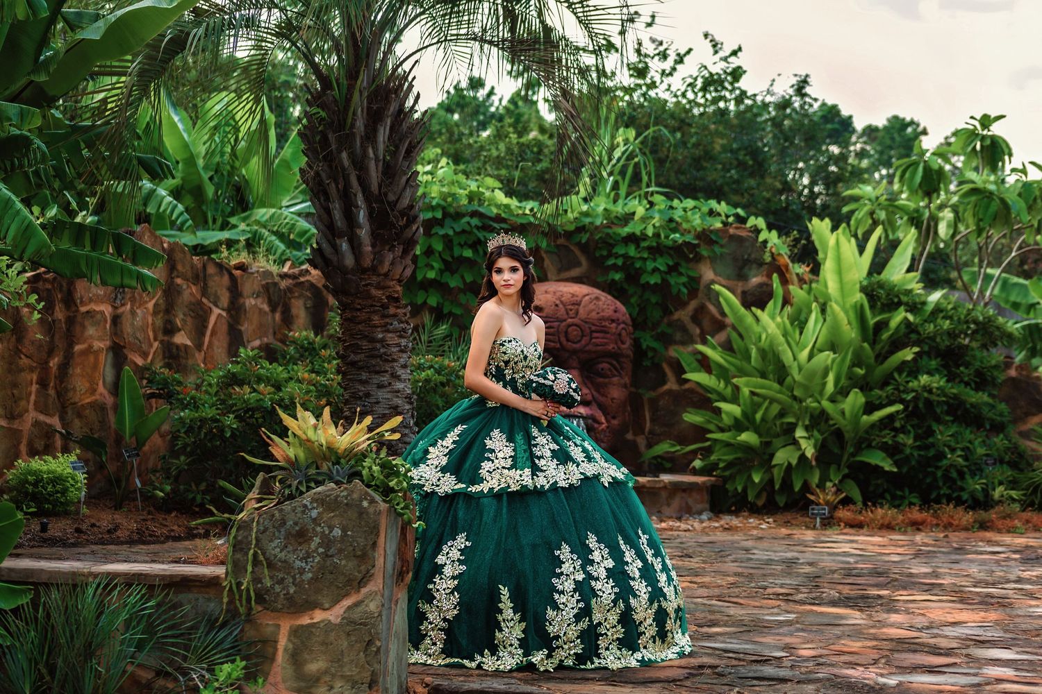 Portrait of Quinceañera girl dressed in green and gold gown standing in park setting