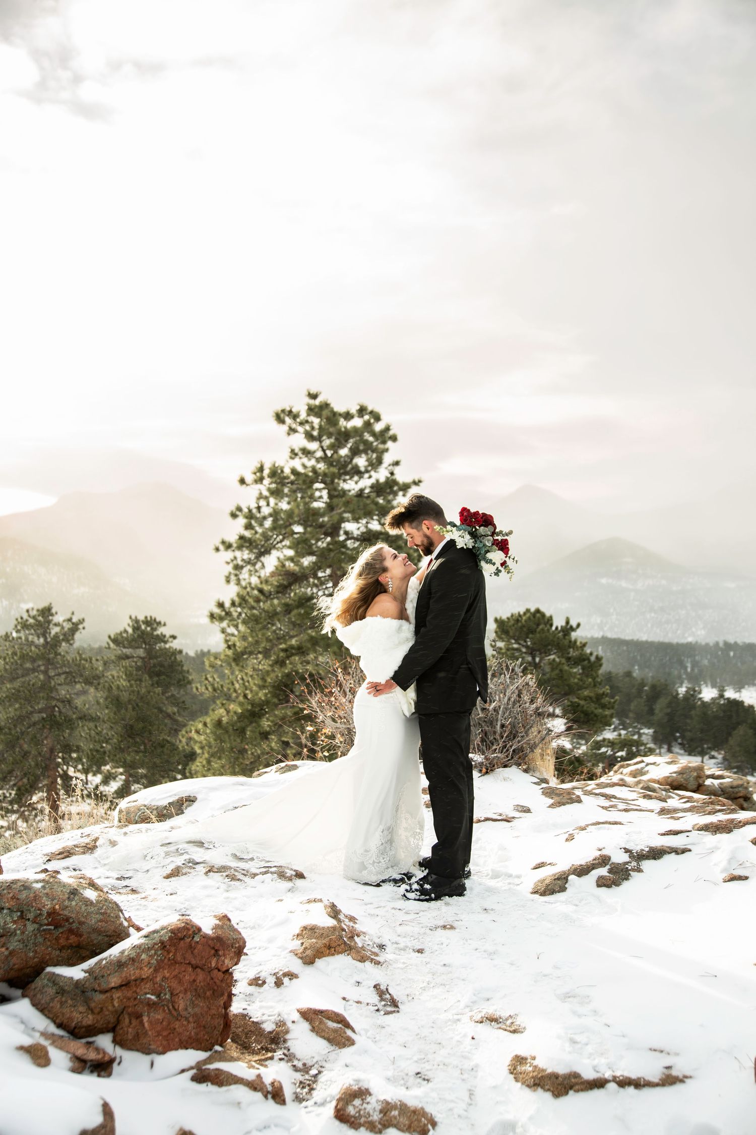 A couple shares an intimate moment on a snowy mountain overlook with evergreen trees and distant peaks in view.