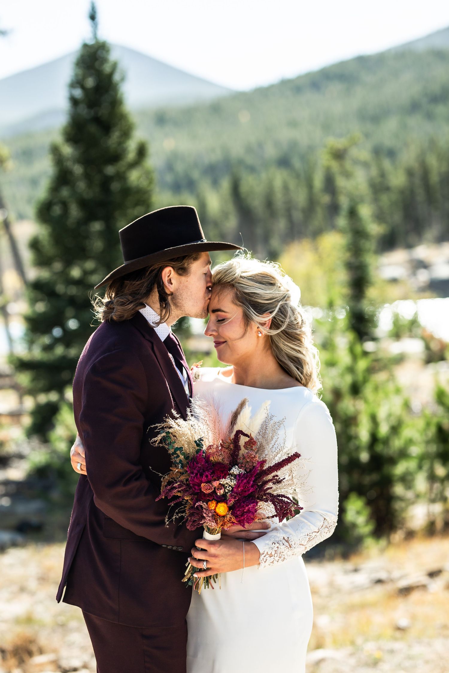 A romantic mountain wedding photo features a couple embracing outdoors with a vibrant burgundy and orange bouquet.