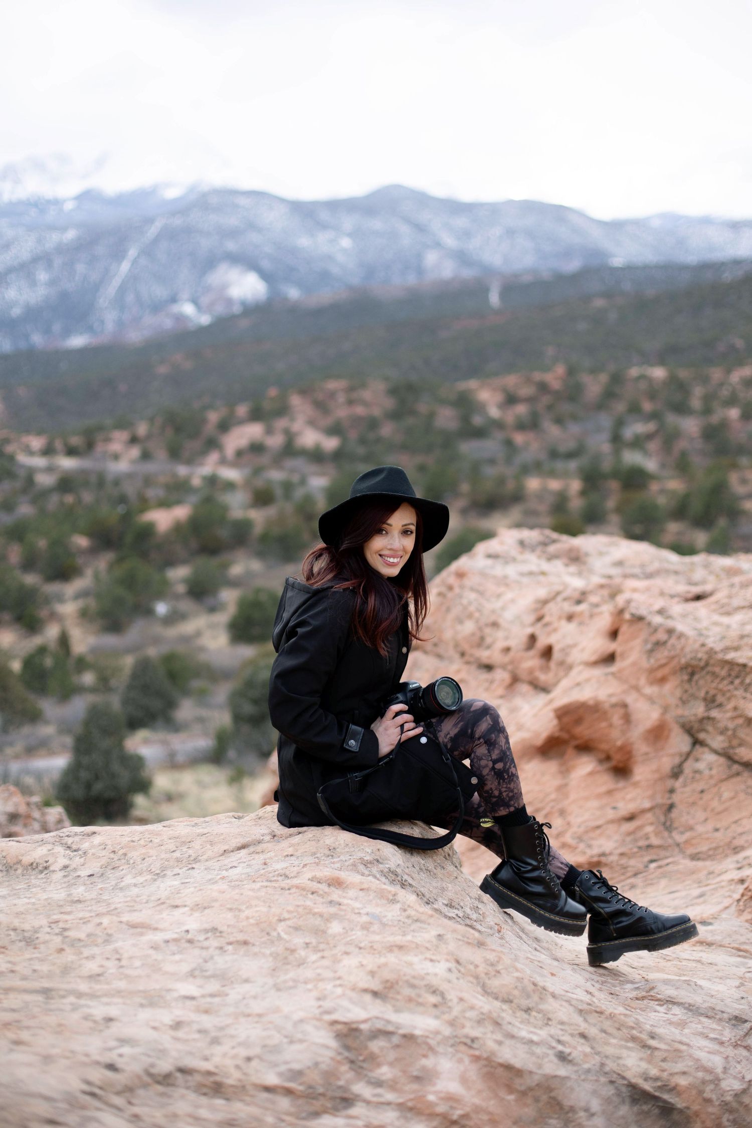 Woman in black sits on red rock cliff edge overlooking snowy mountain landscape.