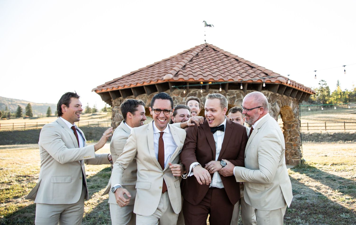 Groomsmen in light colored suits share a laugh together outside a rustic gazebo.