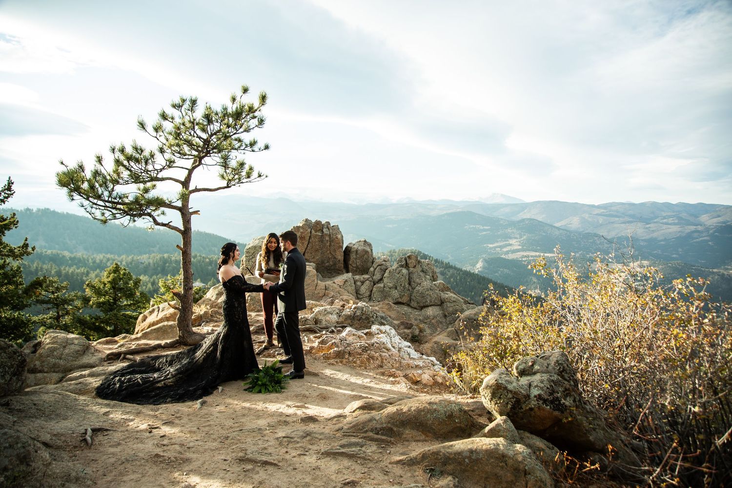 Two people in formal wear stand near a lone pine tree on a rocky mountain overlook with panoramic mountain views.