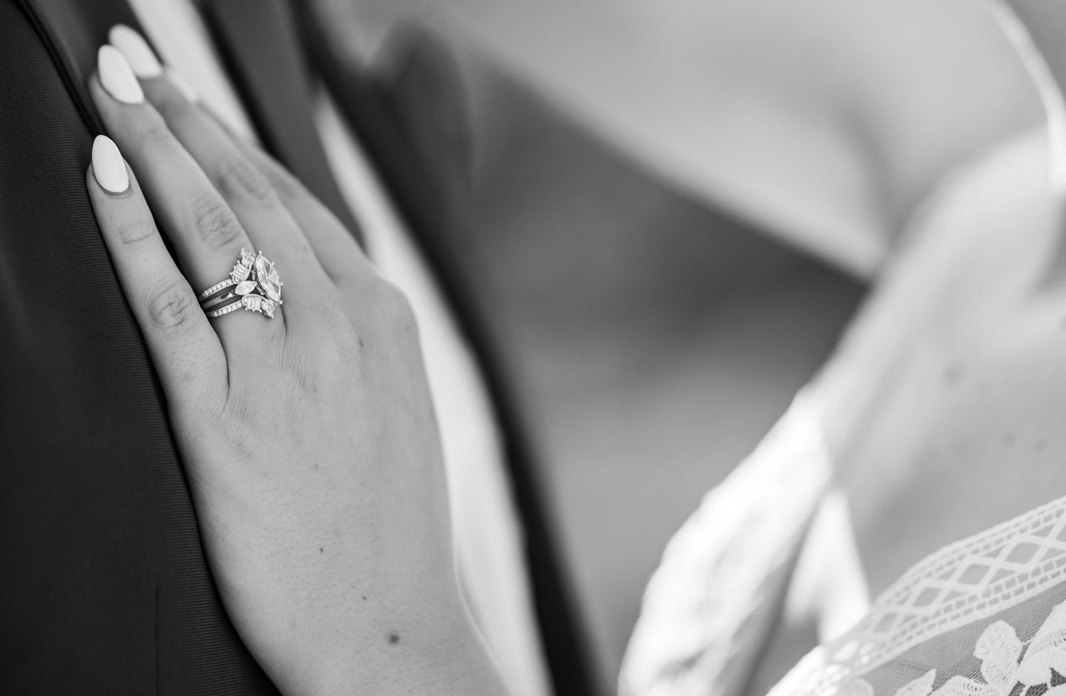A close-up black and white photograph showing an engagement ring on a hand against formal wedding attire.