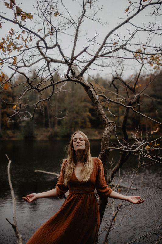 Artistic autumn portrait by lake with bare branches framing figure in rust-colored dress.
