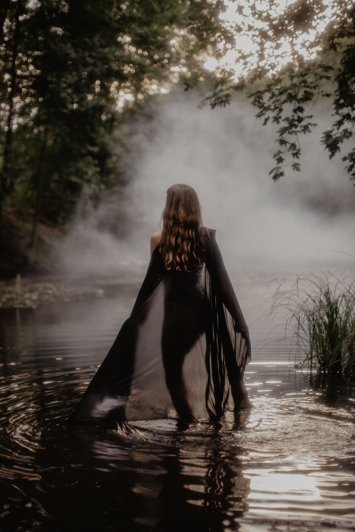 Mysterious figure in black clothing standing in misty water surrounded by forest.