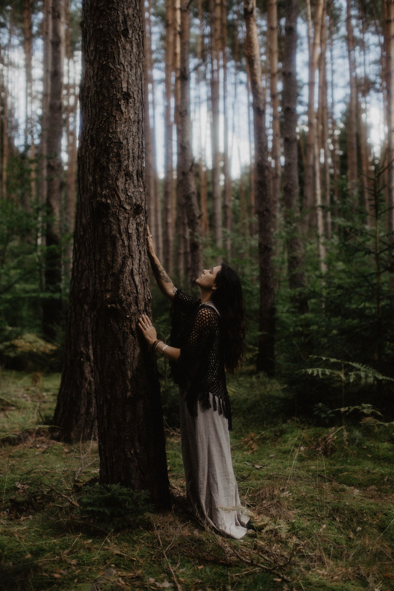 Eine Person in einem langen Kleid steht nachdenklich vor hohen Kiefern in einer nebligen Waldkulisse.