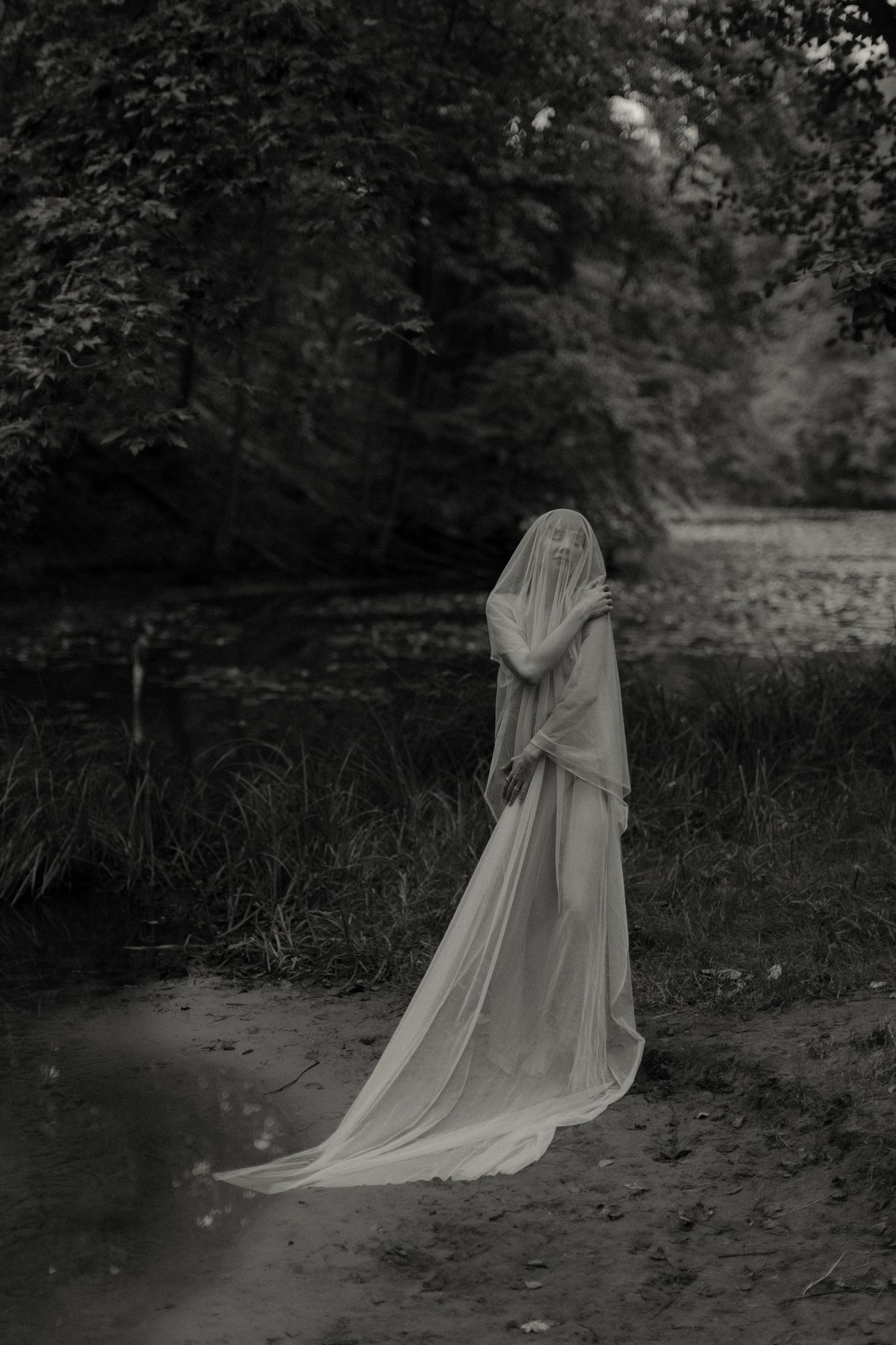 Ethereal black and white photo of a veiled figure in a flowing dress standing in nature.