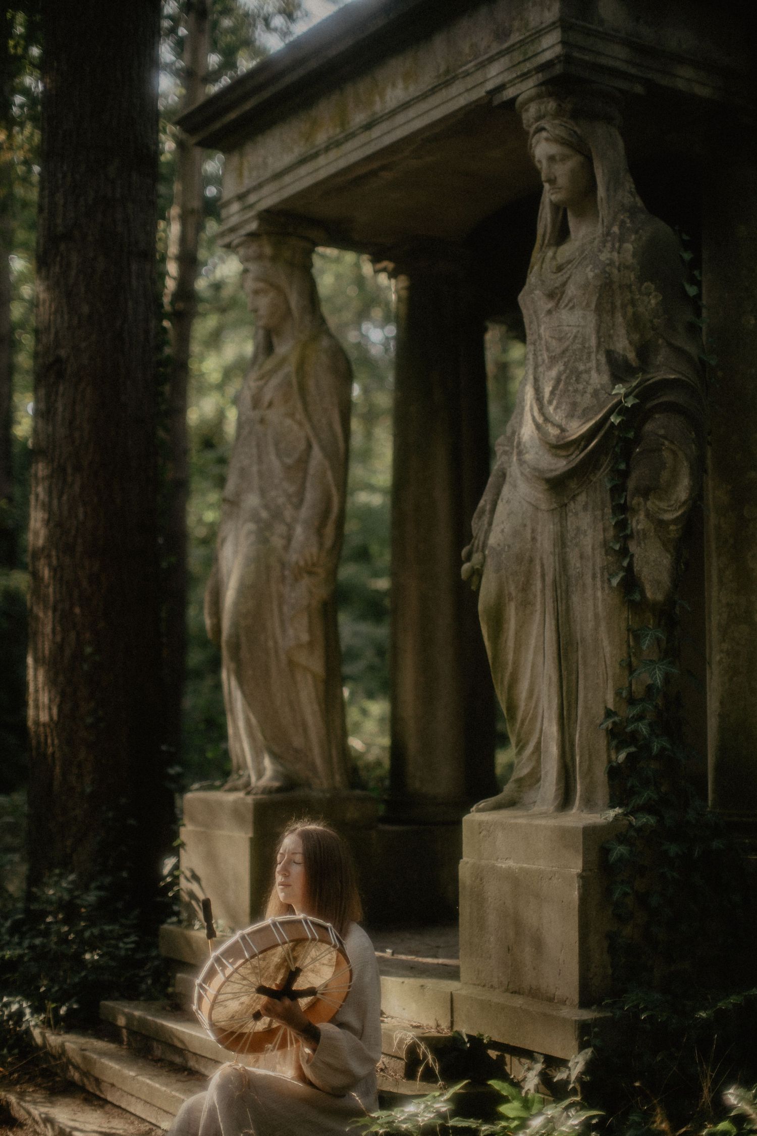 Moody atmospheric photo of vintage stone statues and a seated figure in the forest.