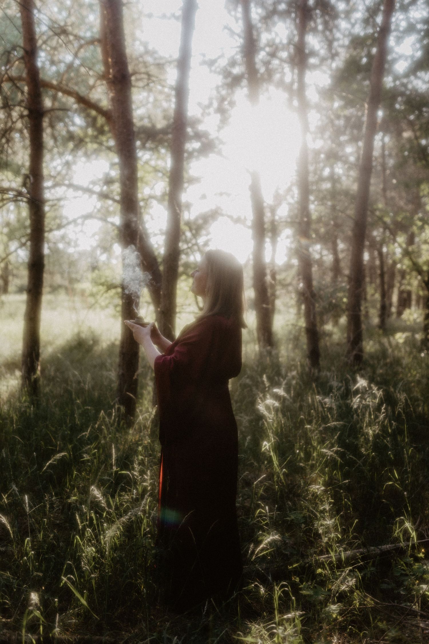 Woman in dark red dress holding smoking object in sunlit forest clearing with tall trees and wild grass.