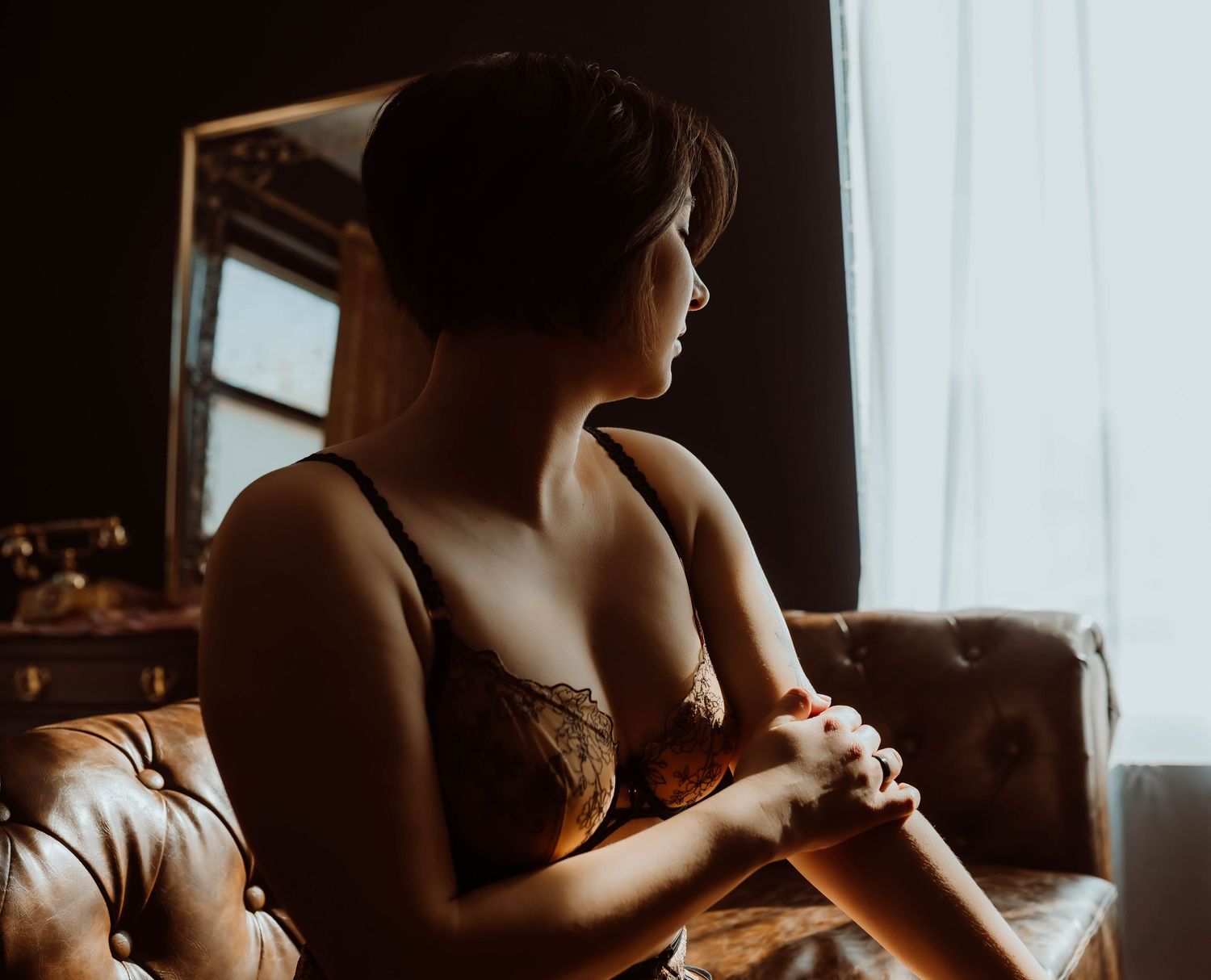A moody, silhouetted figure sits by a window in warm sunlight creating an atmospheric boudoir scene.