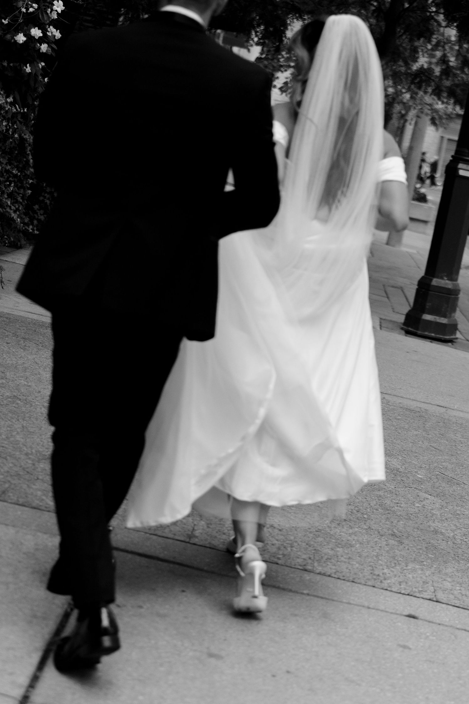 Black and white photo of a bride in flowing white dress and groom in dark suit walking together on a sidewalk.