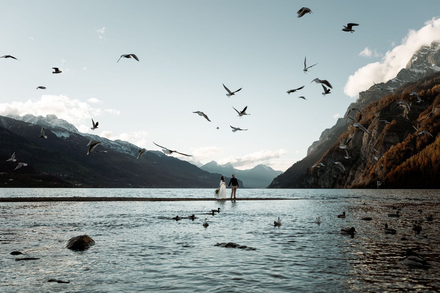 Silhouette figures stand at mountain lake with birds flying overhead against dramatic alpine landscape at dusk.