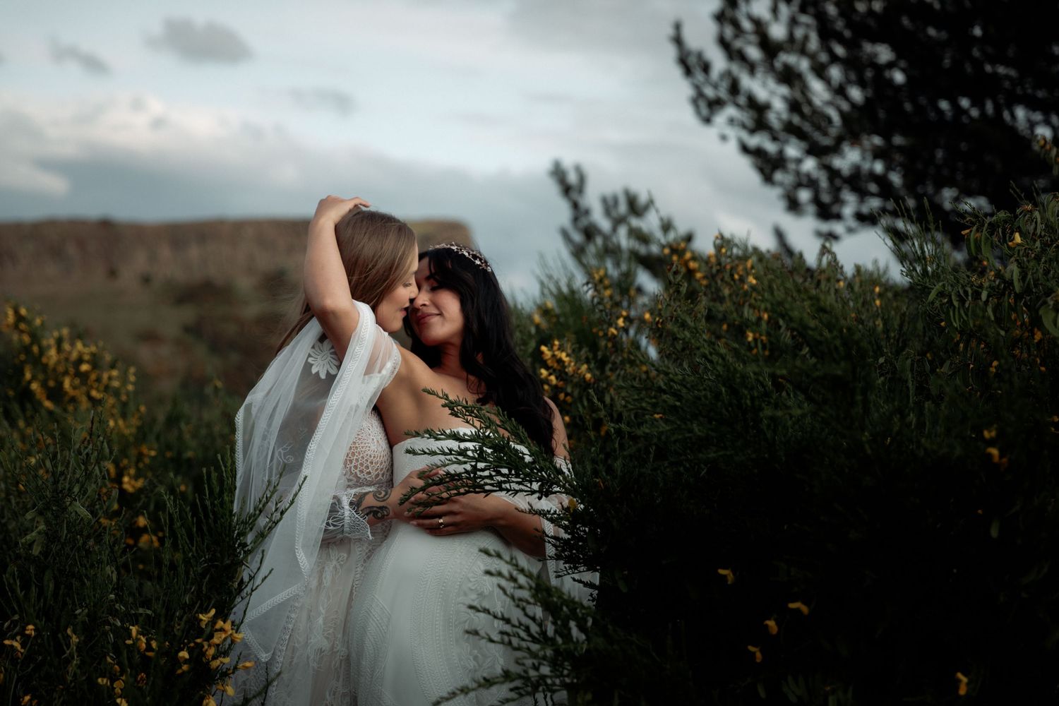 A couple in wedding attire embraces lovingly in a scenic mountain landscape against a cloudy sky at sunset.