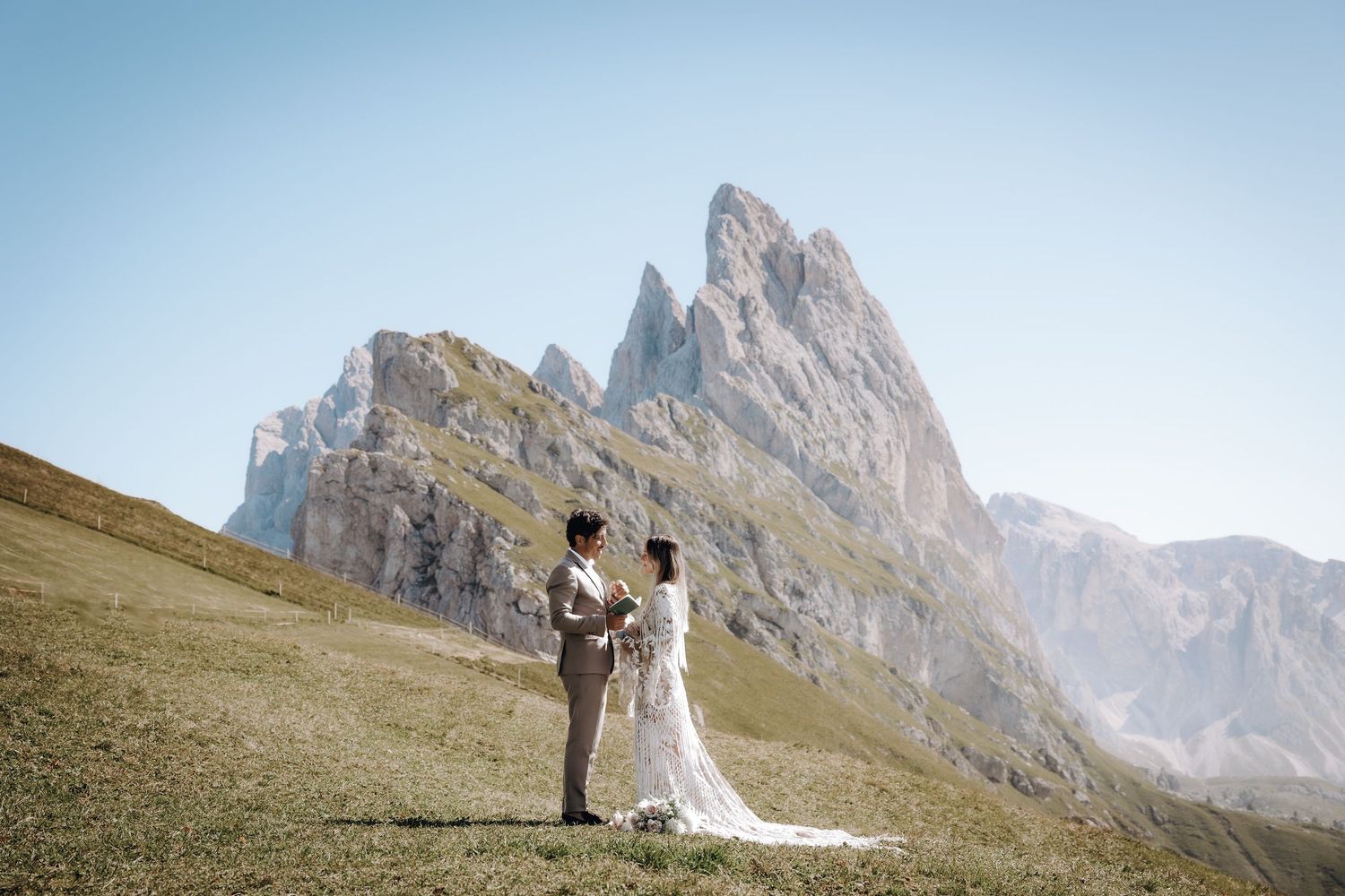 A couple in wedding attire stands on a scenic mountain slope with dramatic rocky peaks in the background.