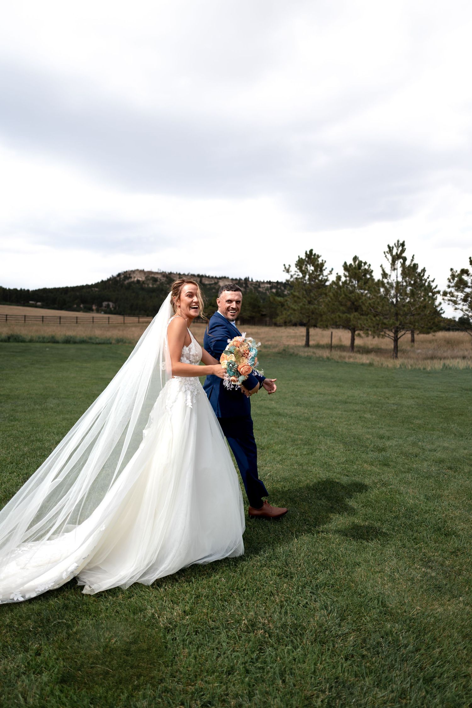 Bride in flowing white gown and veil holds bouquet beside groom in navy suit on green lawn with trees and hills.