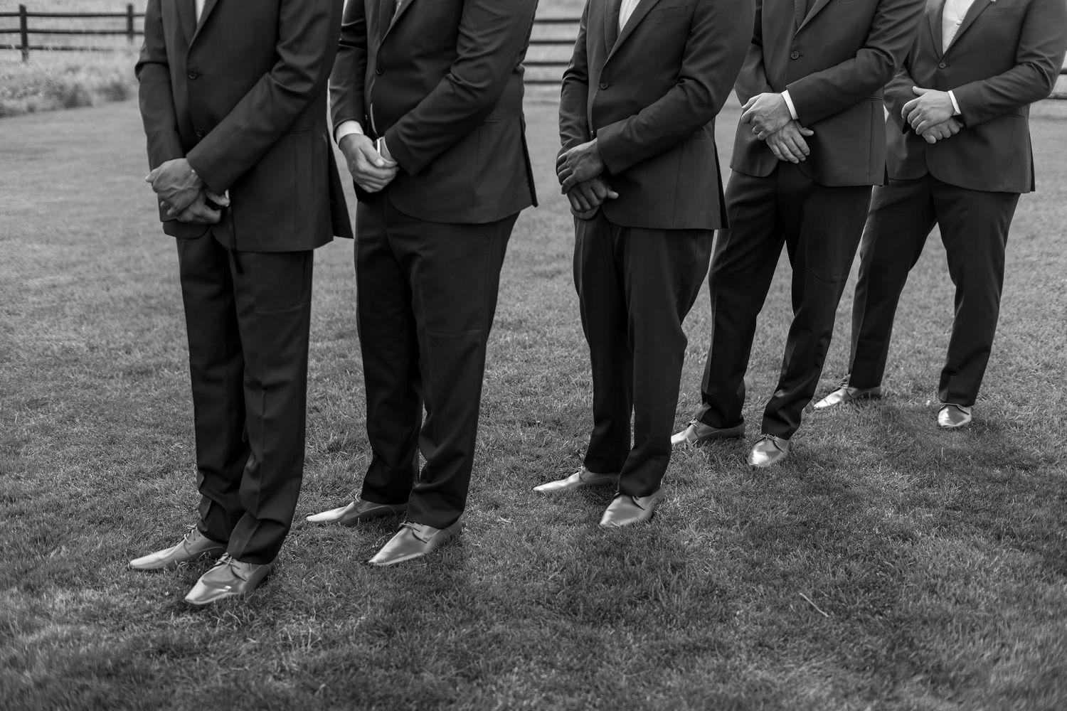 Black and white photo of groomsmen in dark suits standing in a row on grass, showing their metallic dress shoes.